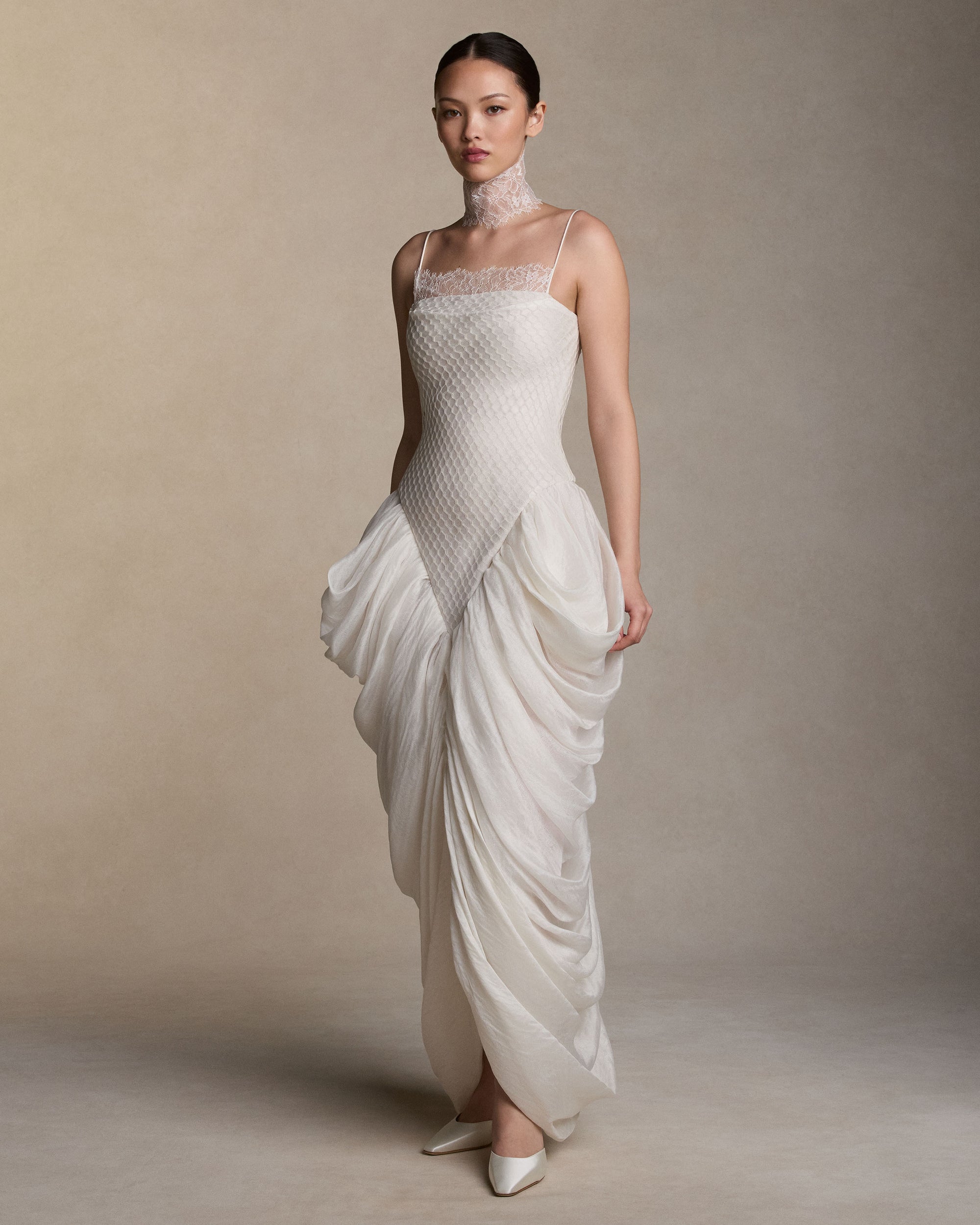 Spaghetti strap gown with a honeycomb lace layered over a silk and wool gauze elongated bodice, with delicate chantilly lace peeking from behind the neckline. Cascading draping falls from the waist creating a tulip skirt silhouette. Internal grosgrain waist belt, invisible zip at center back. 