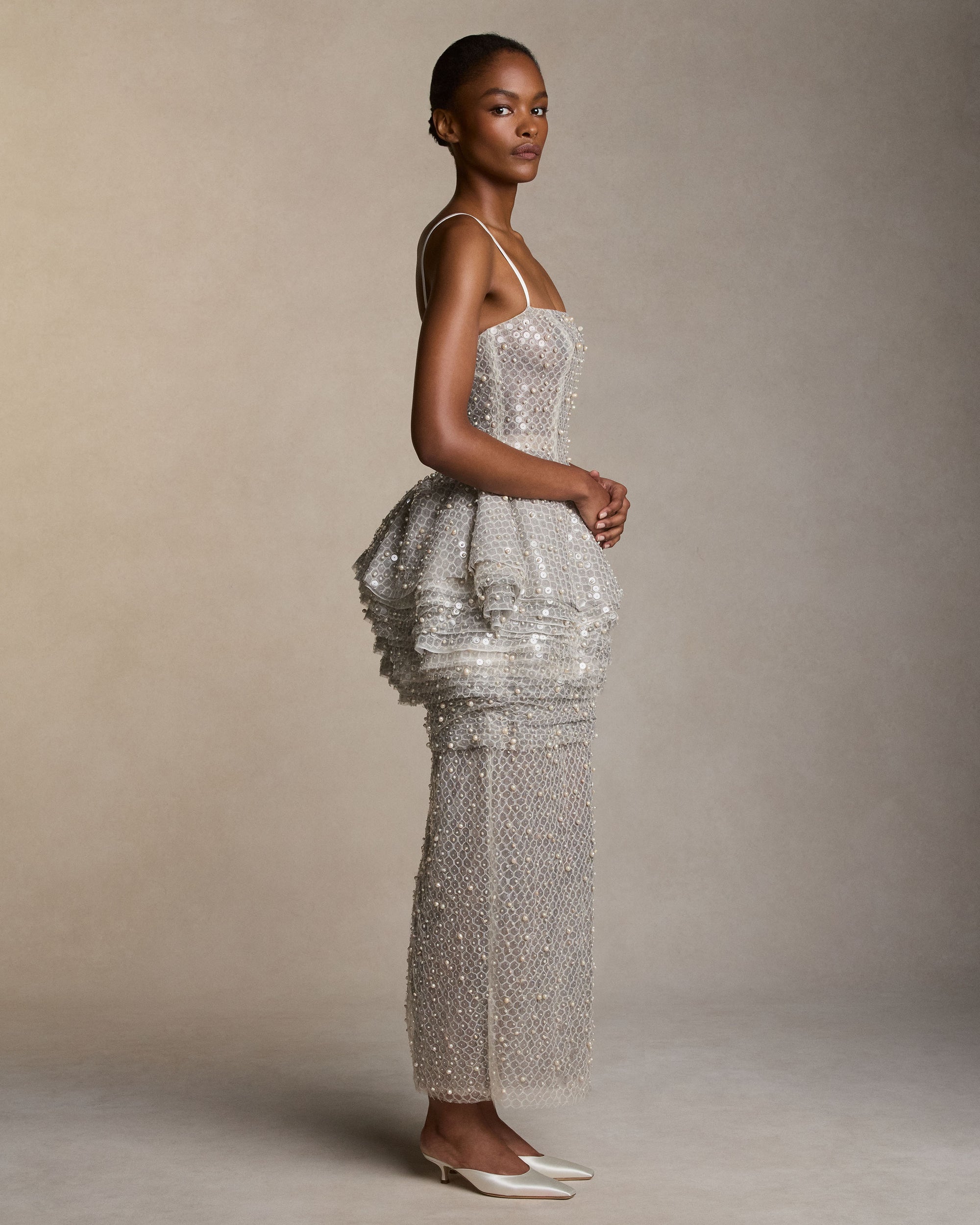 Pegged column gown with tiered peplum skirt and delicate spaghetti straps. Cascading layers of soft tulle, silk organza, and honeycomb lace are delicately hand-embroidered with genuine baroque pearls and transparent sequins. Soft internal bobbinete bustier, grosgrain waistbelt, and invisible zip closure at center back. 