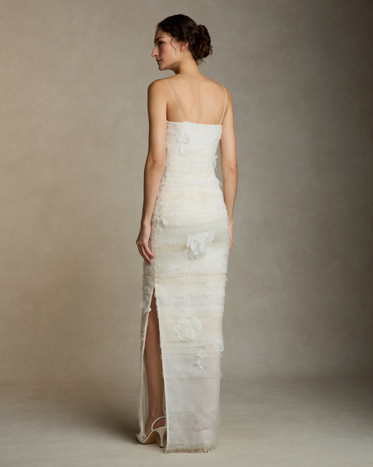 Layered hand cut and frayed organza column dress suspended by delicate spaghetti straps. Hand cut corded and chantilly laces emerge from a gradient of ivory organza strips. Left side slit. Boned internal bustier with side zip closure.Styled with Clara Tulle with Lace Appliqué topper