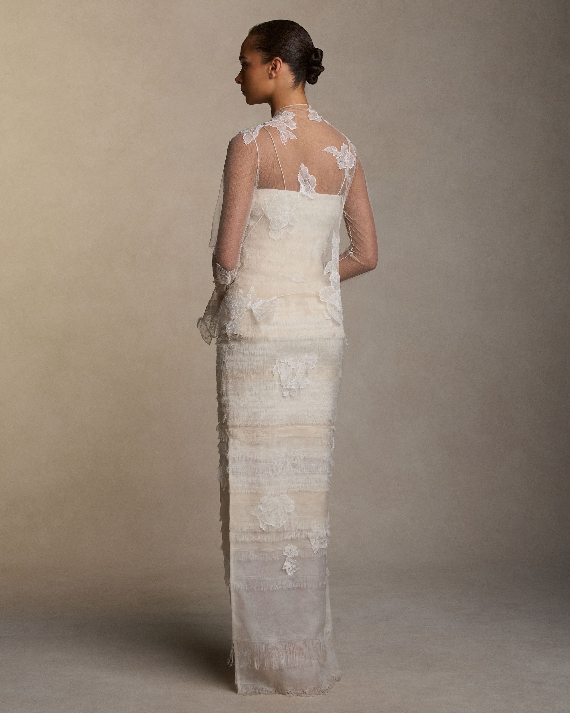Layered hand cut and frayed organza column dress suspended by delicate spaghetti straps. Hand cut corded and chantilly laces emerge from a gradient of ivory organza strips. Left side slit. Boned internal bustier with side zip closure.Styled with Clara Tulle with Lace Appliqué topper
