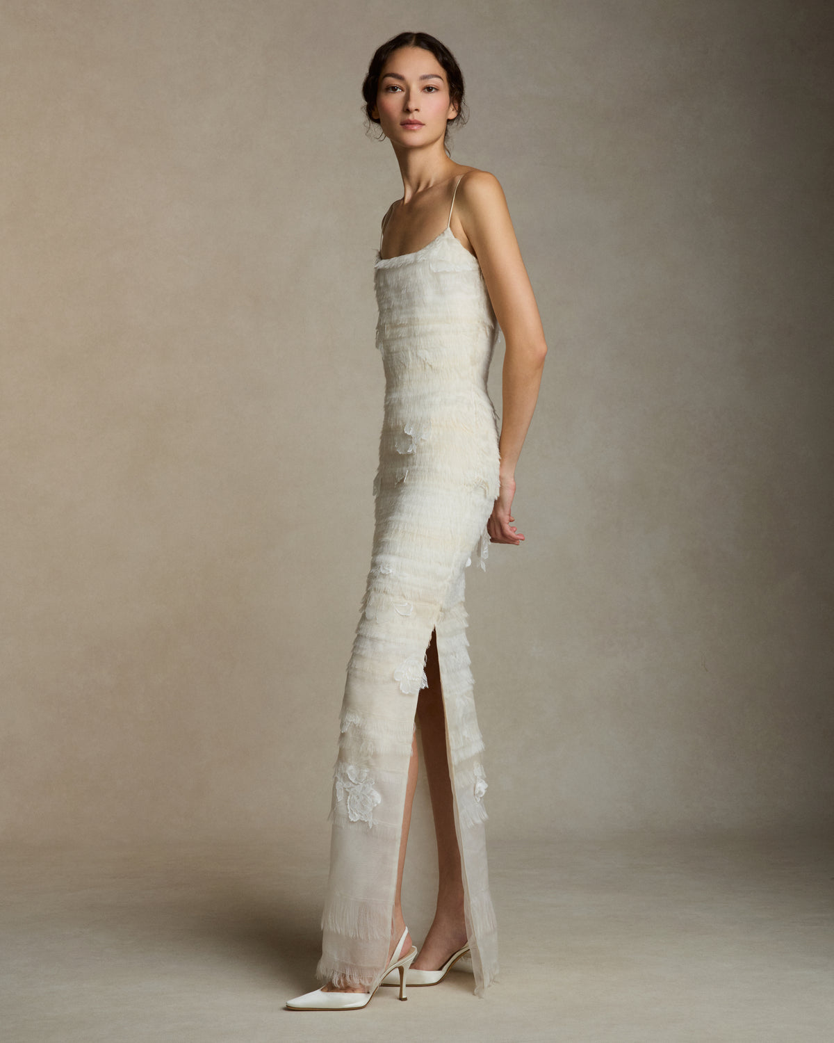 Layered hand cut and frayed organza column dress suspended by delicate spaghetti straps. Hand cut corded and chantilly laces emerge from a gradient of ivory organza strips. Left side slit. Boned internal bustier with side zip closure.Styled with Clara Tulle with Lace Appliqué topper