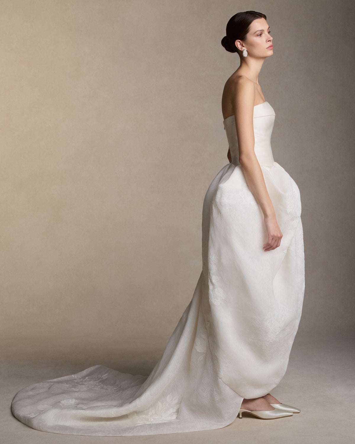 Silk wool twill and linen organza strapless ball gown. Structured silk wool twill bodice is finished with a foldover neckline. A draped linen organza skirt, decorated with hand painted florals and corded and Chantilly lace appliqués, releases into a godet train. Boned internal bustier, internal grosgrain belt and invisible zip closure at center back.