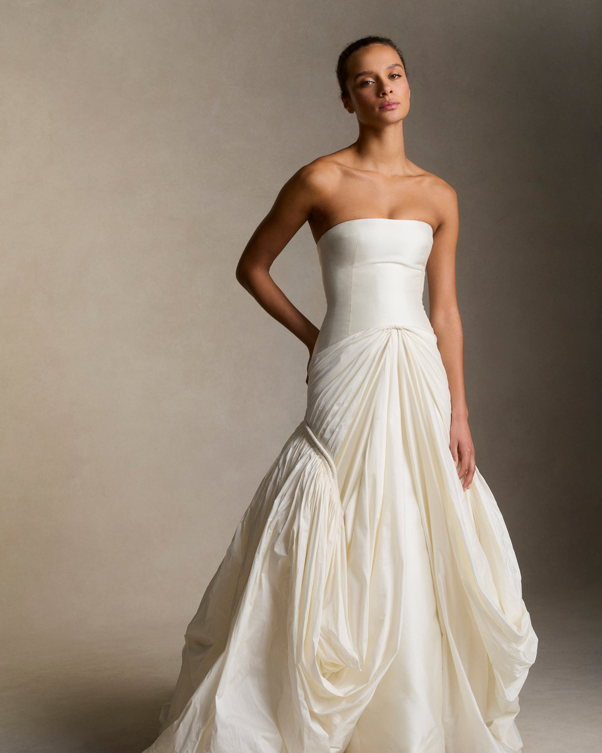 Strapless silk wool twill column gown with draped and gathered tissue taffeta bubble hem skirt. Corded channel detail frames the hip silhouette disappearing into draped pickup detail at center front. Boned internal bustier, internal grosgrain belt and invisible zip closure at center back.