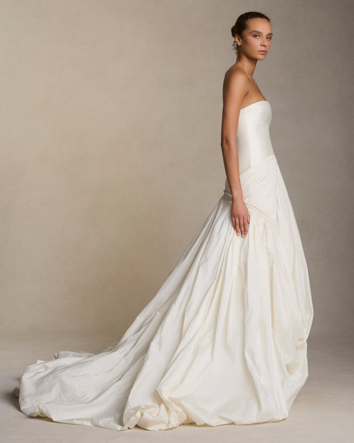 Strapless silk wool twill column gown with draped and gathered tissue taffeta bubble hem skirt. Corded channel detail frames the hip silhouette disappearing into draped pickup detail at center front. Boned internal bustier, internal grosgrain belt and invisible zip closure at center back.