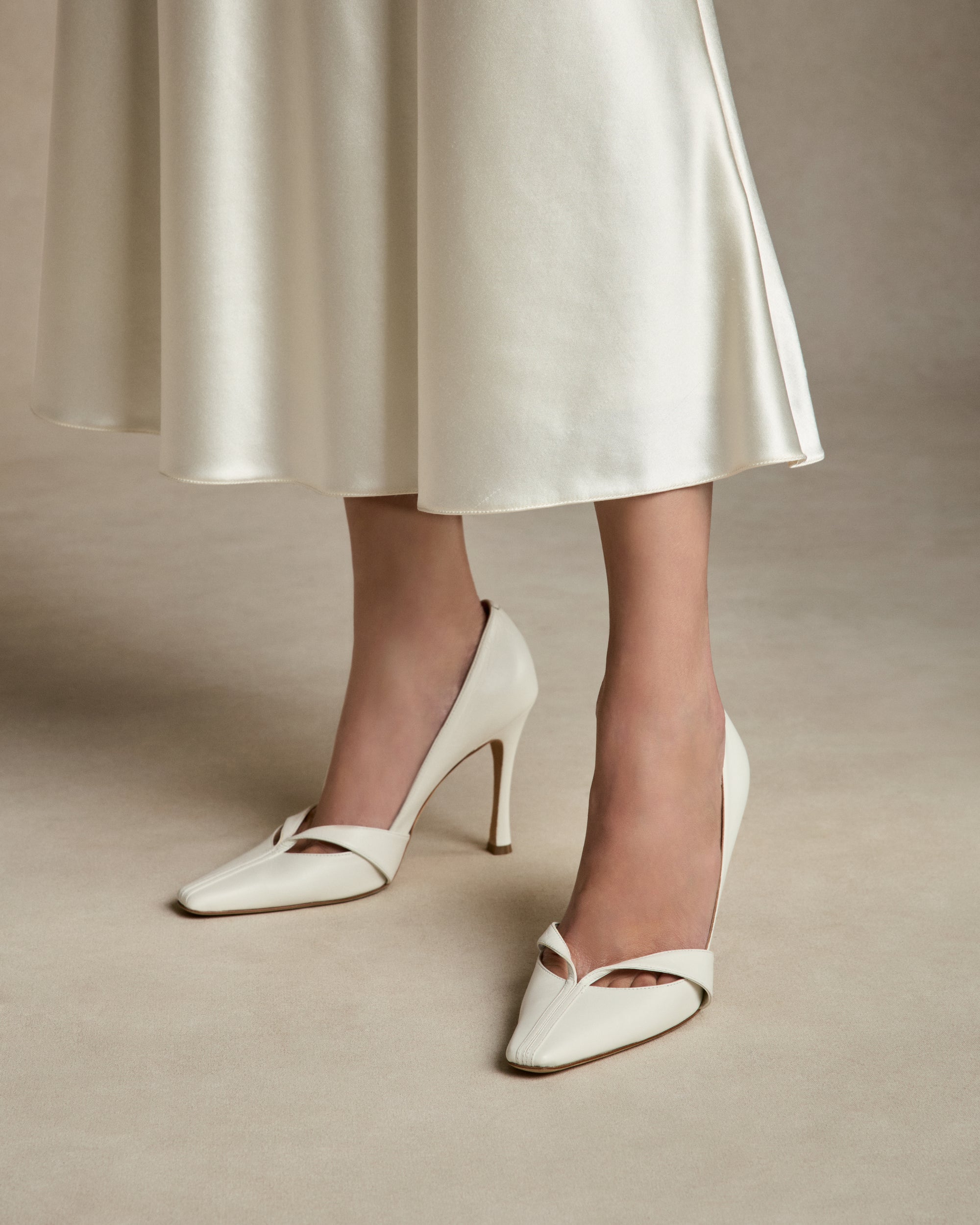 Rounded square toe pump crafted in Italy of ivory leather. Leather trim detail decorates the toe, peeling away at neckline as it wraps around the foot. 100mm heel.