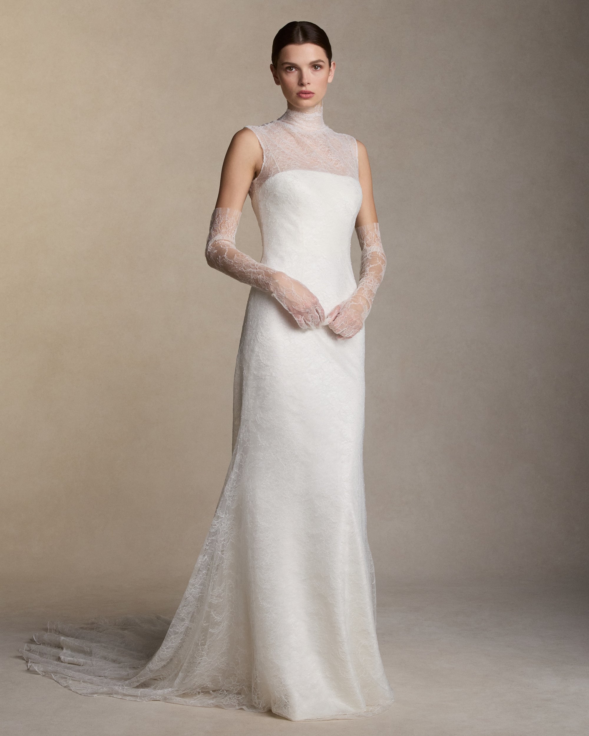 High neck sleeveless chantilly lace gown with covered button closure at center back. Layers of delicate chantilly lace finish in scalloped edge detail at neck with self fabric covered buttons down center back. Soft internal bustier with invisible zip closure at center back.