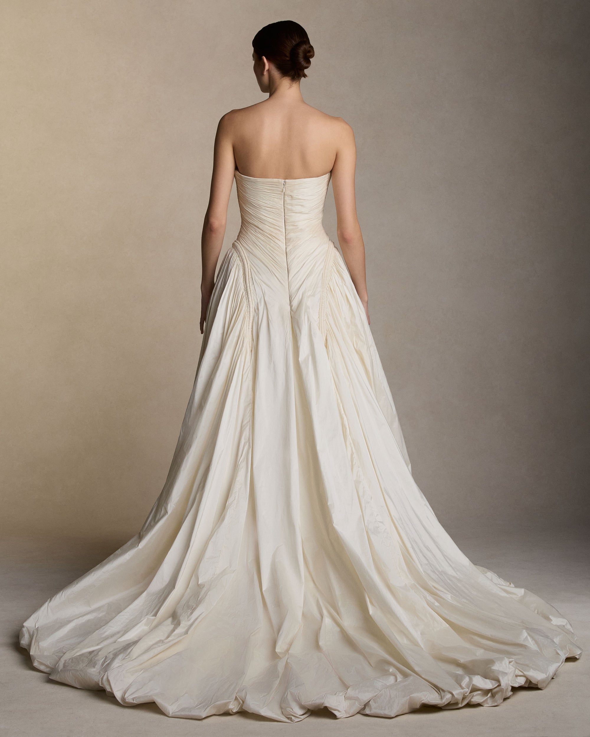 Strapless tissue taffeta ballgown with soft dipped neckline. Hand pleated peaked bust draping releases from a center front twist. Corded channel details frame the bodice silhouette and transition into a bubbled ball skirt. Boned internal bustier, internal grosgrain belt and invisible zip closure at center back. Styled with Pleated Overlay