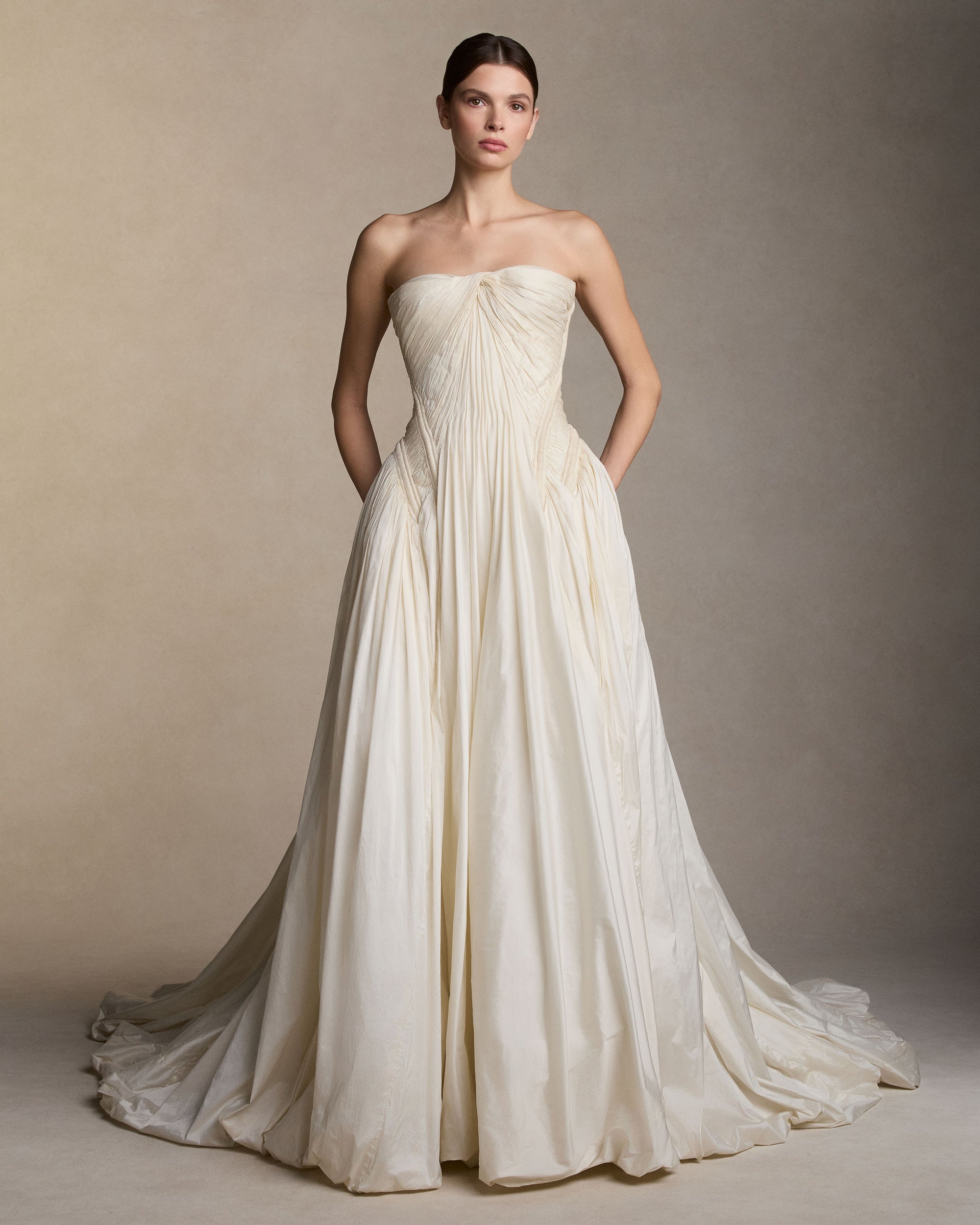 Strapless tissue taffeta ballgown with soft dipped neckline. Hand pleated peaked bust draping releases from a center front twist. Corded channel details frame the bodice silhouette and transition into a bubbled ball skirt. Boned internal bustier, internal grosgrain belt and invisible zip closure at center back. Styled with Pleated Overlay