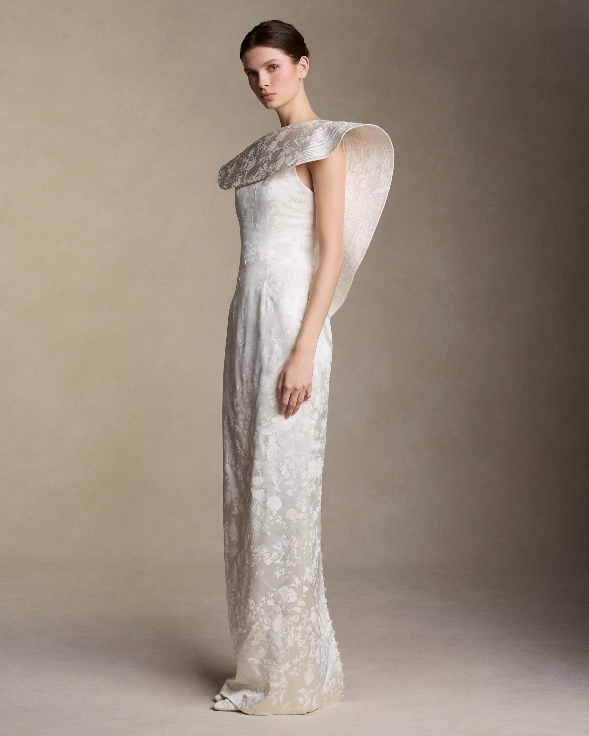 Floral pigment printed and hand painted silk wool column gown. Exaggerated fluted collar drape with trapunto stitch detailing peels from neckline falling down center back. Internal grosgrain belt and invisible zip closure at center back.