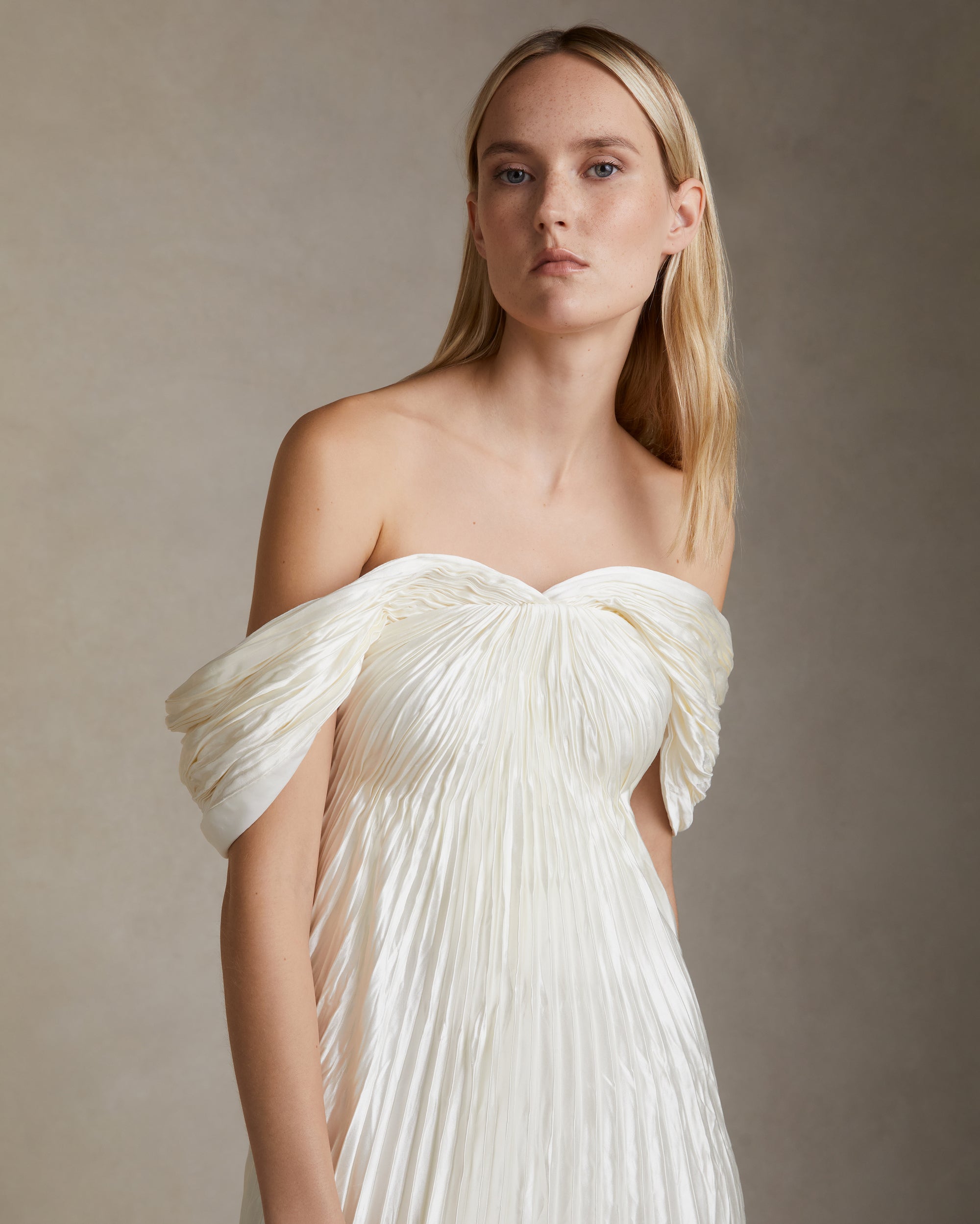 Off the shoulder pleated and hand crinkled silk wool gown with swagged sleeve drape releasing from center front neckline. Boned internal bustier, soft cups, internal grosgrain waist belt and invisible center back zip closure. 
