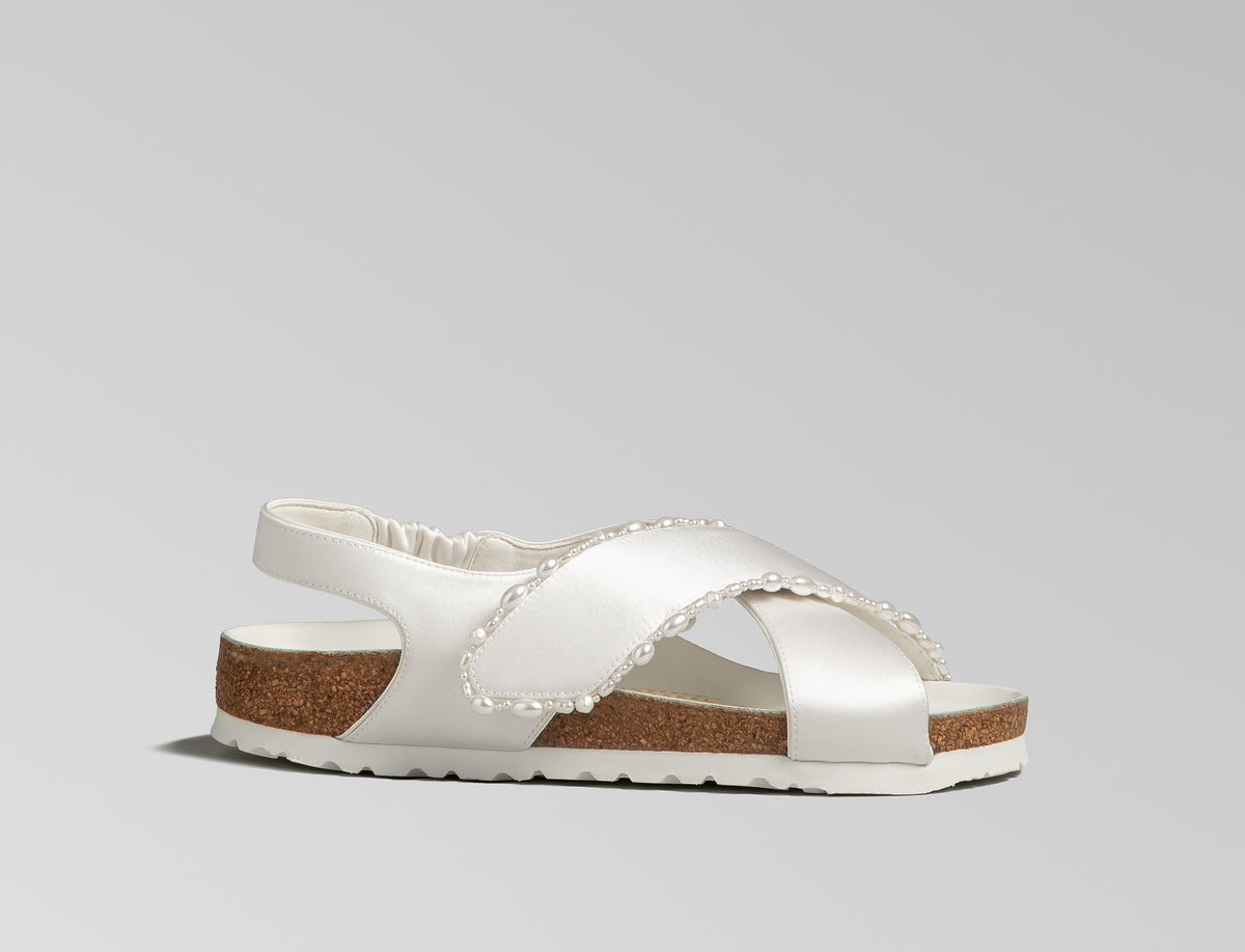 The Tulum is reimagined in a luminous ivory satin, featuring a graceful crossover profile and a slim ankle strap adorned with delicate pearls. The design also introduces an innovative adjustable hidden buckle closure. Handcrafted in Germany, the shoe merges BIRKENSTOCK’s legendary comfort with Danielle Frankel’s signature touch of refined elegance.- Upper: Delicate satin upper, naturally shaped pearls handstitched on trim- Footbed: Anatomical shaped BIRKENSTOCK cork latex Footbed lined with premium Nappa Leather- Sole: EVA die cut outsole with a custom design, blending classic BIRKENSTOCK bone pattern with delicate leaf inspired detailing
