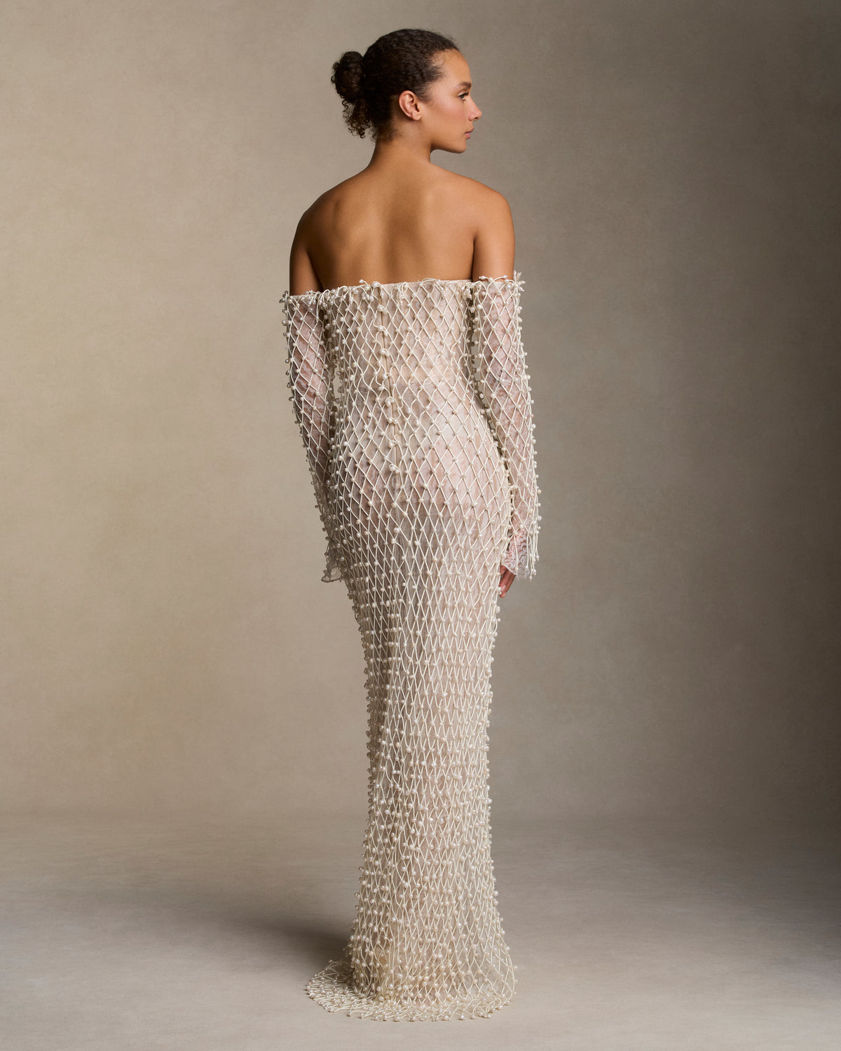 Warped transparent organza off the shoulder column gown with hand applied corded trellis embroidery embellished in transparent sequins and baroque pearls. Fringe hem detail at sleeves is finished with genuine teardrop pearls. Bobinette lining with boned internal bustier. Invisible zip closure at center back.