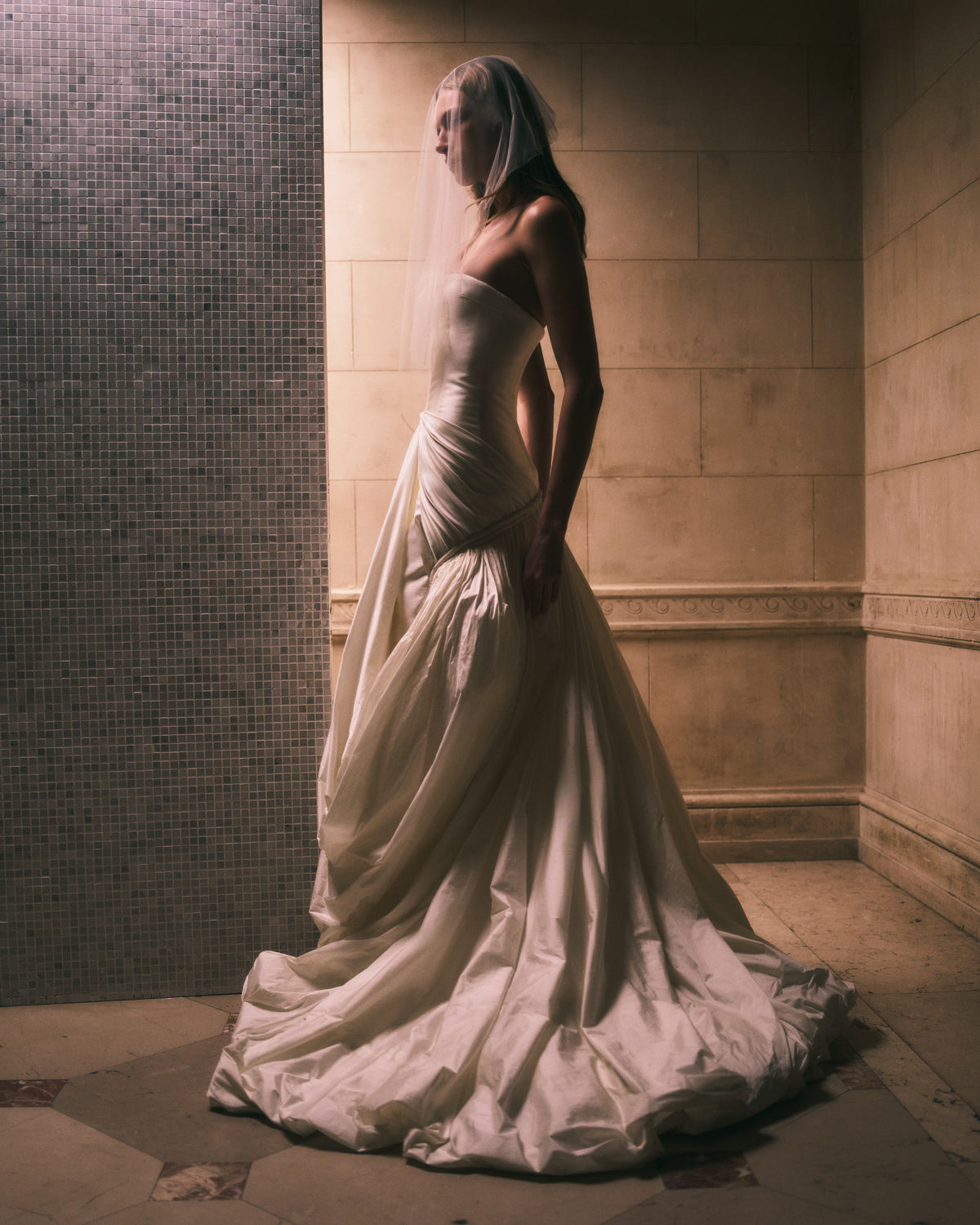 Strapless silk wool twill column gown with draped and gathered tissue taffeta bubble hem skirt. Corded channel detail frames the hip silhouette disappearing into draped pickup detail at center front. Boned internal bustier, internal grosgrain belt and invisible zip closure at center back.