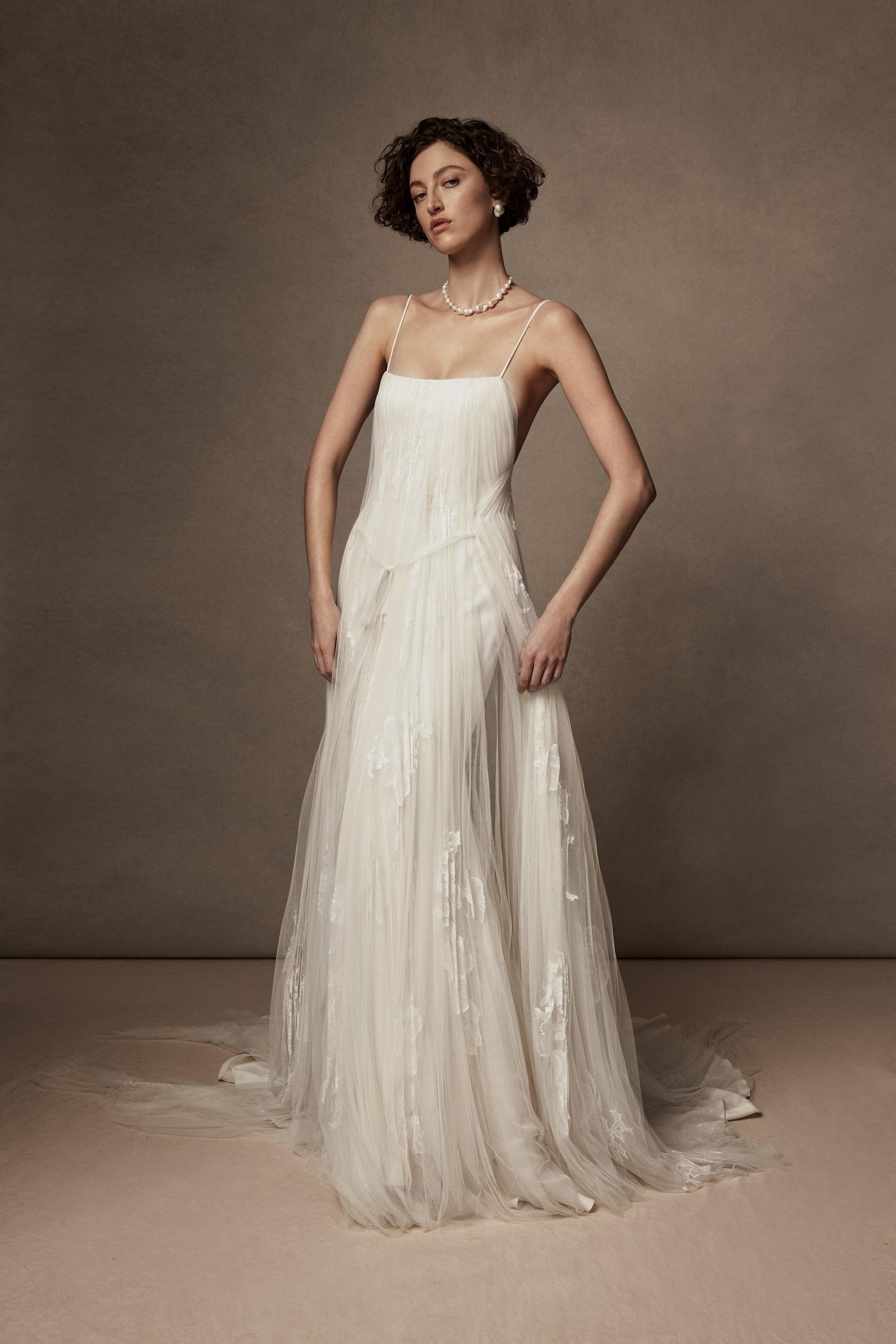 Layered sunburst pleated tulle and lace appliquéd gown. Draping from neckline and flowing through low v-back. Embellished with hand cut corded and chantilly laces. Delicate tulle accent tie at waist. Soft internal bustier with side boning, cups, and grosgrain belt with 3 hook position. Low center back zip, hook and eye closure. Layered and removable tulle shirt with chantilly lace appliqué.