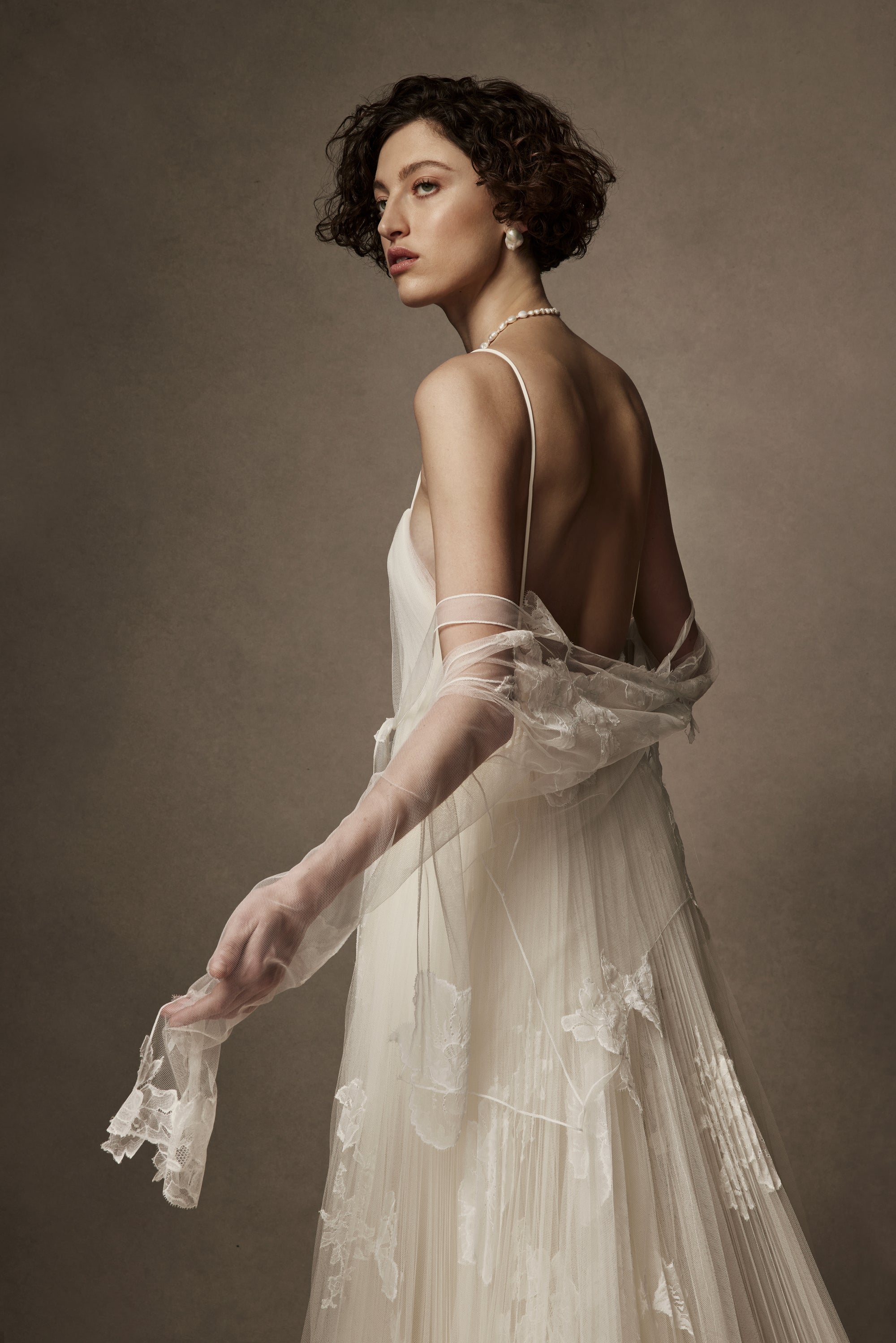 Layered sunburst pleated tulle and lace appliquéd gown. Draping from neckline and flowing through low v-back. Embellished with hand cut corded and chantilly laces. Delicate tulle accent tie at waist. Soft internal bustier with side boning, cups, and grosgrain belt with 3 hook position. Low center back zip, hook and eye closure. Layered and removable tulle shirt with chantilly lace appliqué.