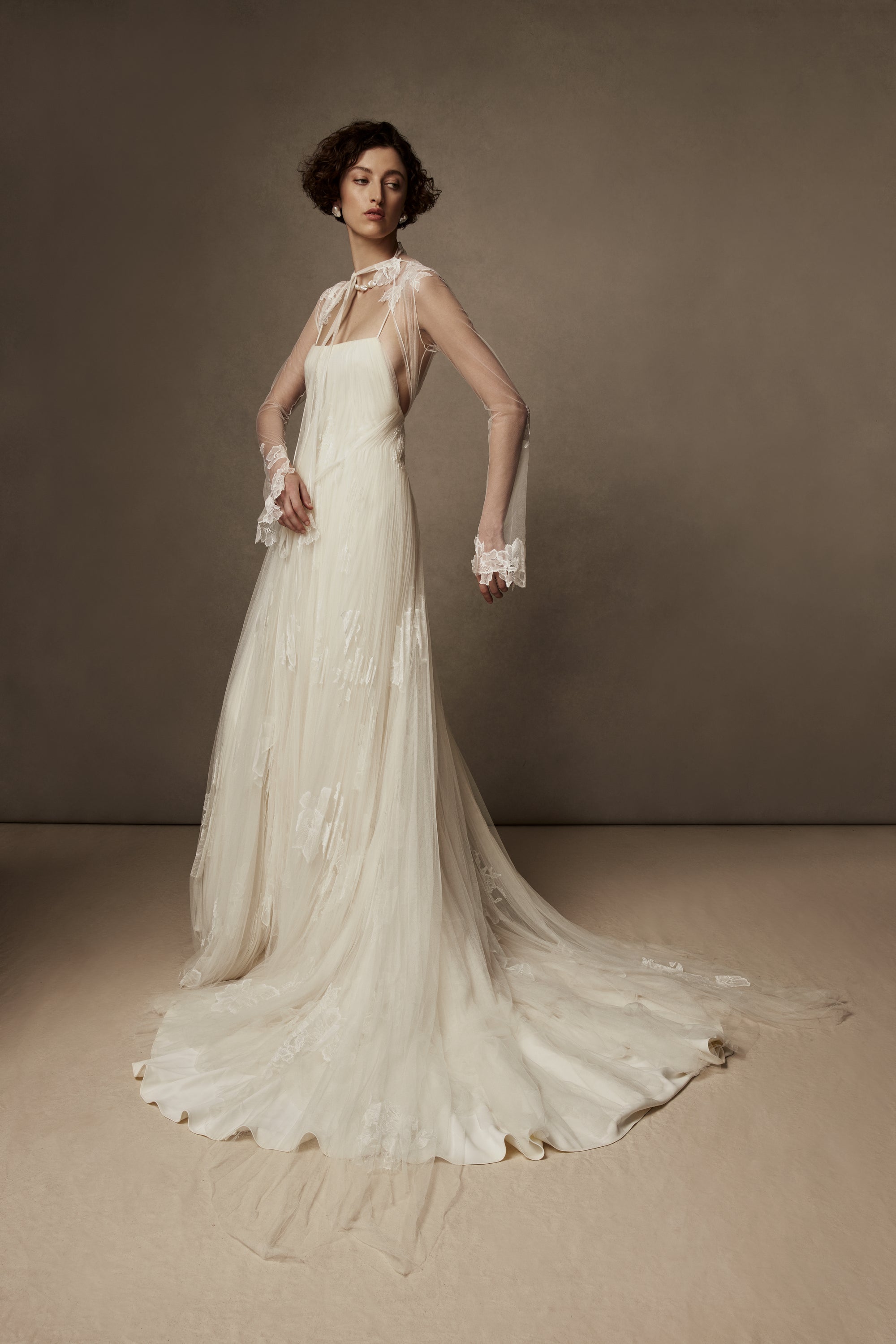 Layered sunburst pleated tulle and lace appliquéd gown. Draping from neckline and flowing through low v-back. Embellished with hand cut corded and chantilly laces. Delicate tulle accent tie at waist. Soft internal bustier with side boning, cups, and grosgrain belt with 3 hook position. Low center back zip, hook and eye closure. Layered and removable tulle shirt with chantilly lace appliqué.