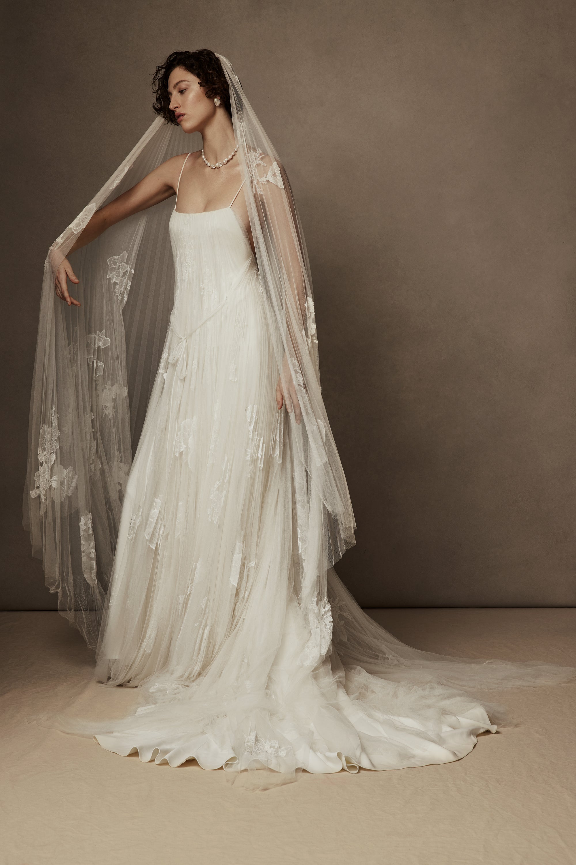Layered sunburst pleated tulle and lace appliquéd gown. Draping from neckline and flowing through low v-back. Embellished with hand cut corded and chantilly laces. Delicate tulle accent tie at waist. Soft internal bustier with side boning, cups, and grosgrain belt with 3 hook position. Low center back zip, hook and eye closure. Layered and removable tulle shirt with chantilly lace appliqué.