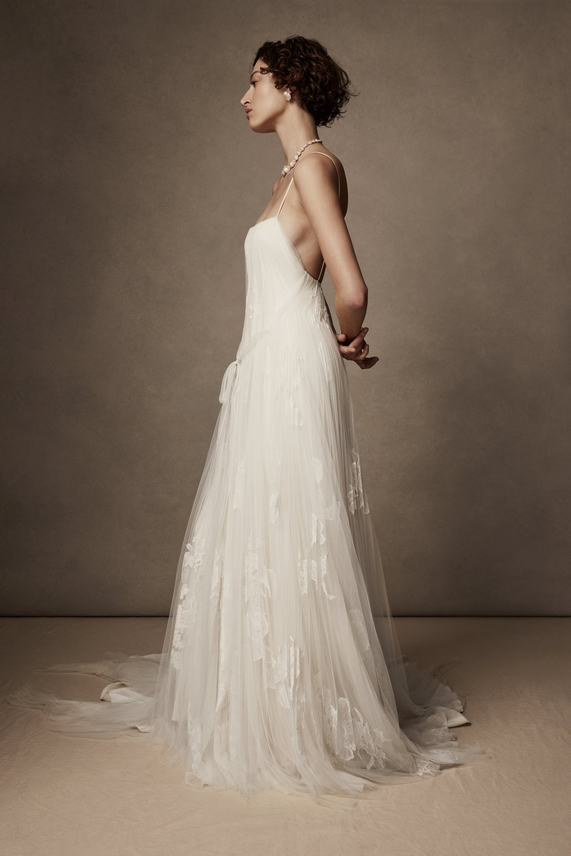 Layered sunburst pleated tulle and lace appliquéd gown. Draping from neckline and flowing through low v-back. Embellished with hand cut corded and chantilly laces. Delicate tulle accent tie at waist. Soft internal bustier with side boning, cups, and grosgrain belt with 3 hook position. Low center back zip, hook and eye closure. Layered and removable tulle shirt with chantilly lace appliqué.