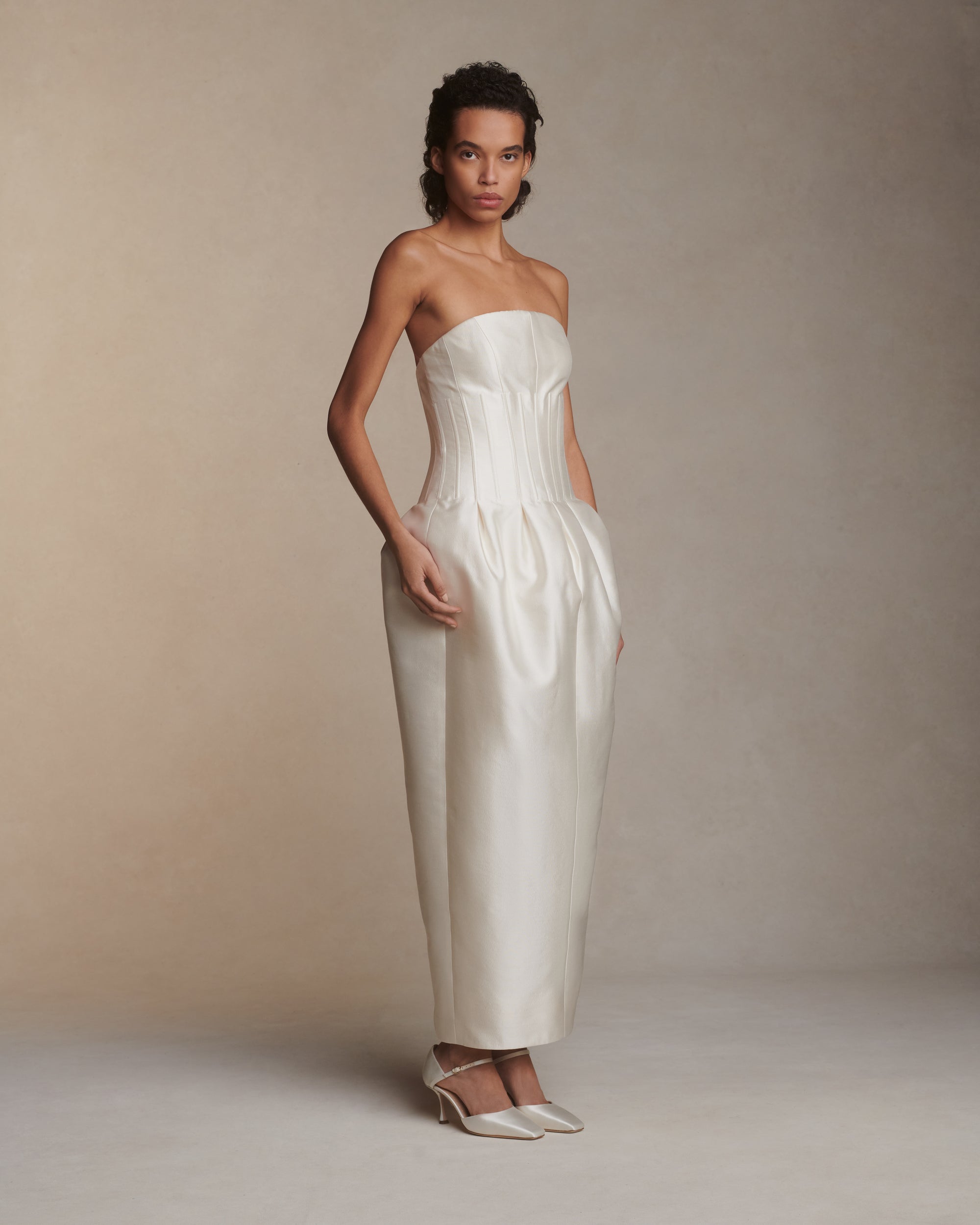 Luminous silk wool twill tea length gown. Boned waist with sculptural inverted pleats extends into a structured bust with curved neckline. Side seam pockets, soft cups, internal grosgrain waistband, and center back zip closure.