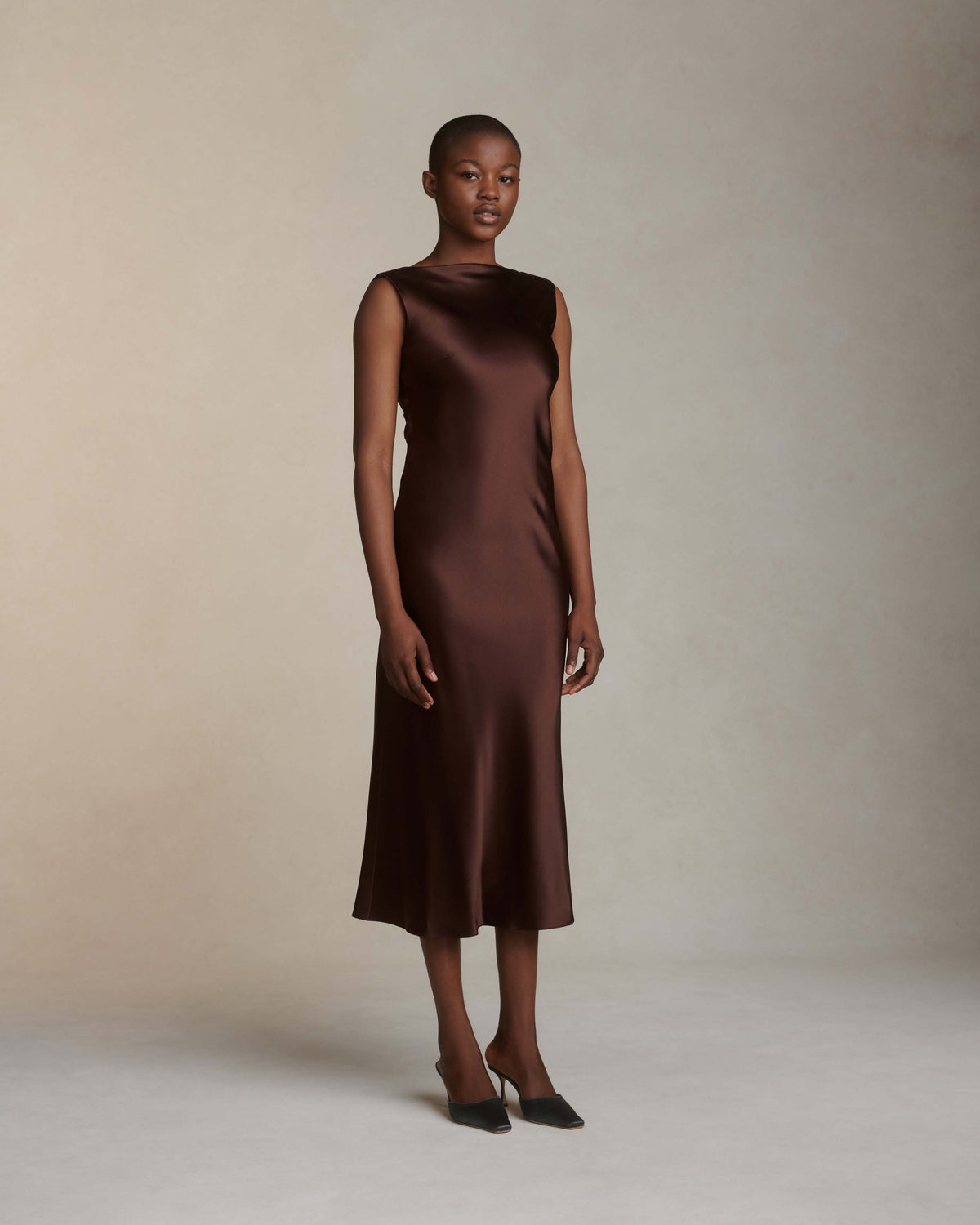 Raised cowl neck, bias cut silk satin sleeveless gown. Open back with scarf draped accent. Fully lined with side zip closure. Midi length.