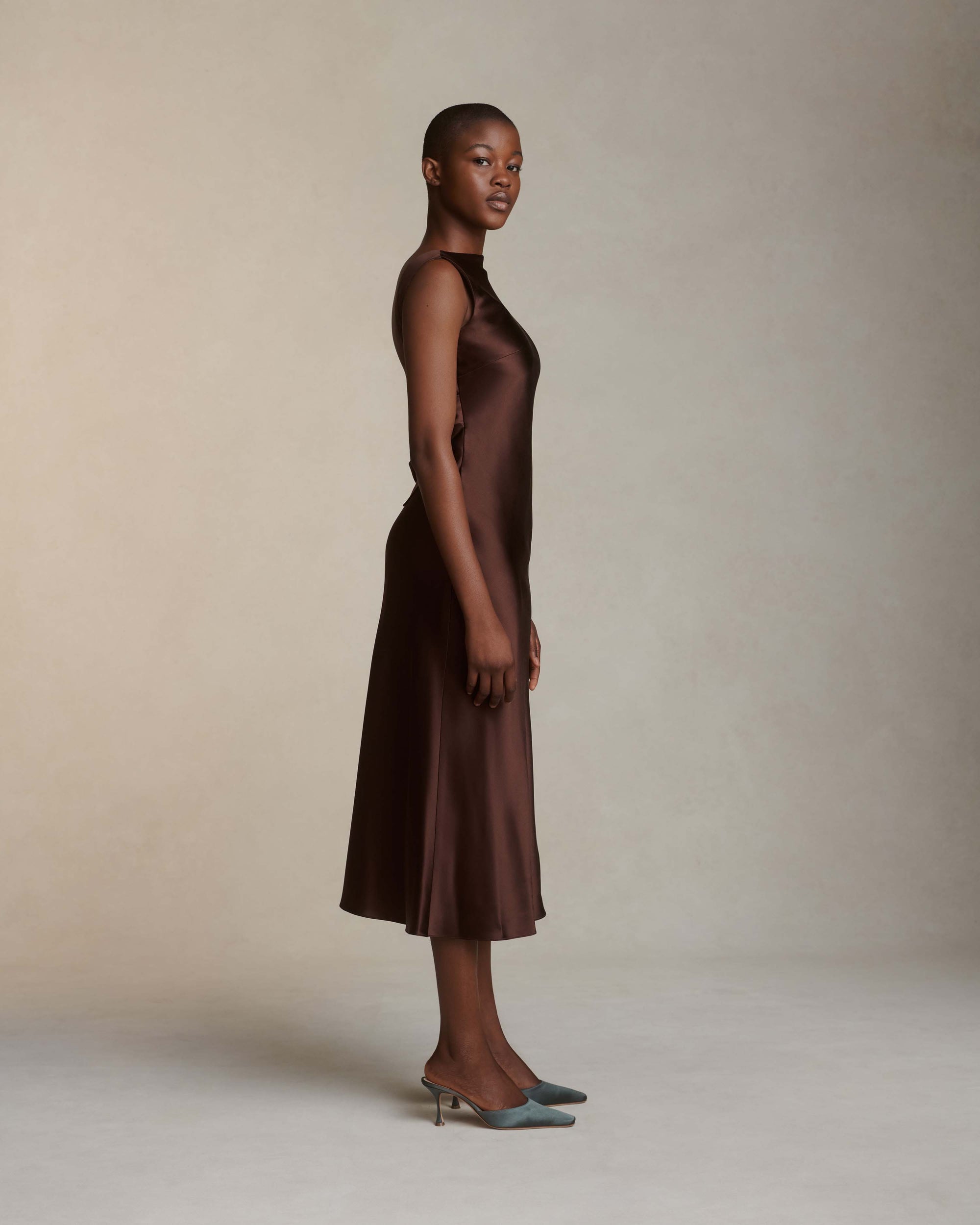 Raised cowl neck, bias cut silk satin sleeveless gown. Open back with scarf draped accent. Fully lined with side zip closure. Midi length.