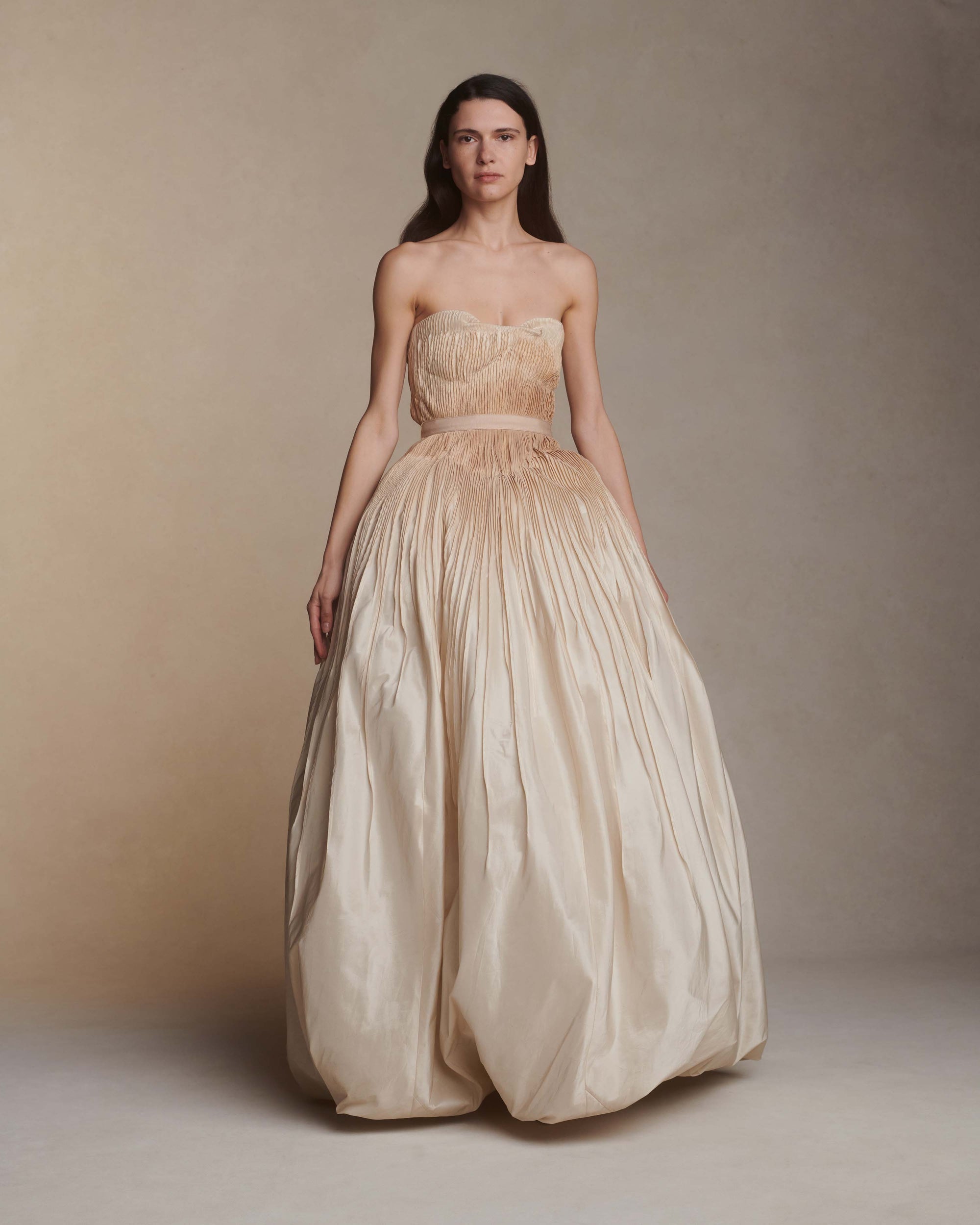 Hand tucked silk taffeta gown with dipped neckline. Micro tucked drop waist bodice with grosgrain waist tie releases into a dramatic skirt with bubbled hem. Hand-dyed ombre bodice fades from under bust and into skirt. Soft internal cups, boning, internal waist belt, invisible center back zip. Available in Ivory $10,990.