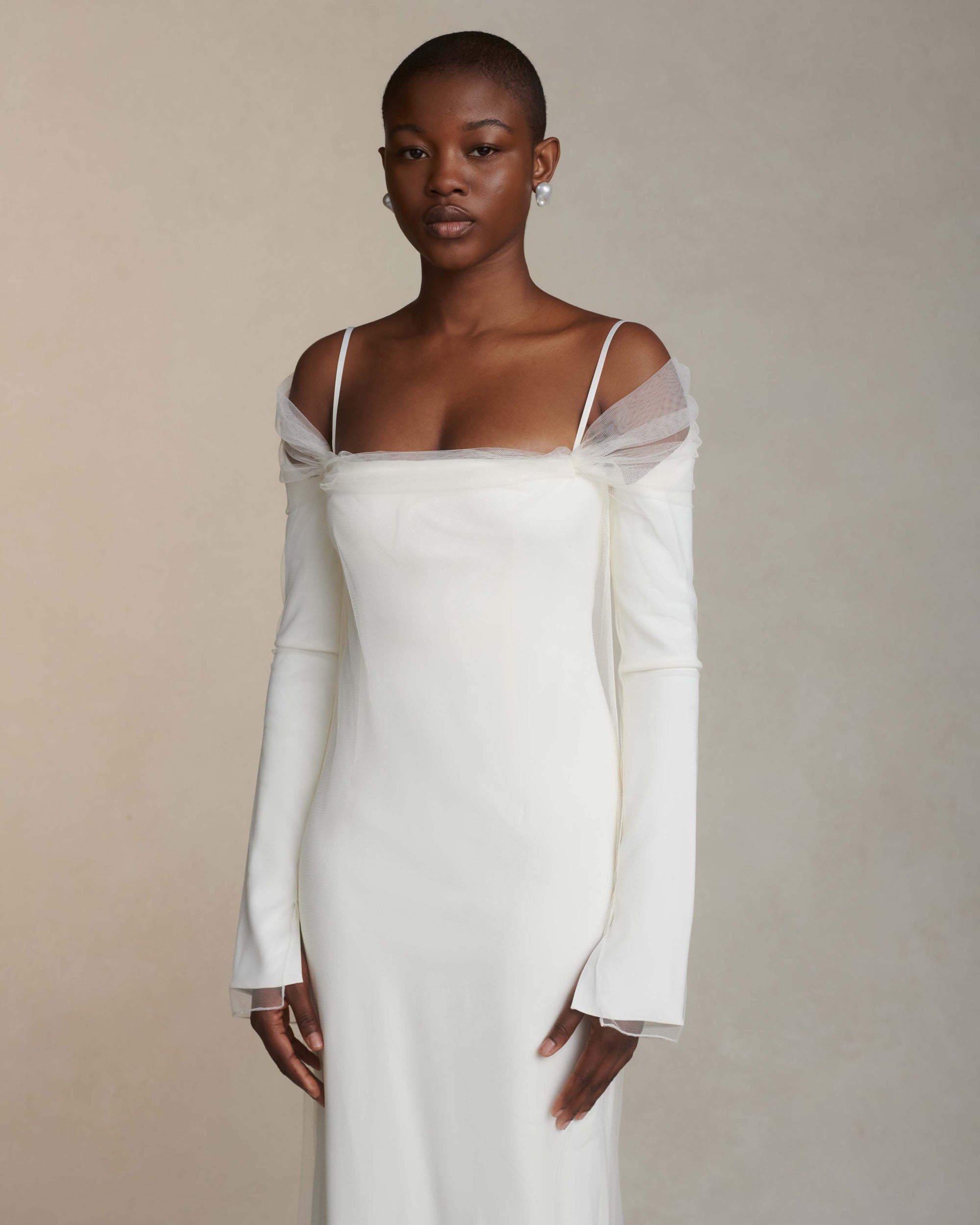 Stretch Cady off the shoulder gown with extended tulle overlay. Squared neckline accented by draped high tulle, long sleeves lightly bound to narrow satin ribbon straps. Center back external zipper and soft internal bustier with grosgrain waistband belt and three hook positions for support.