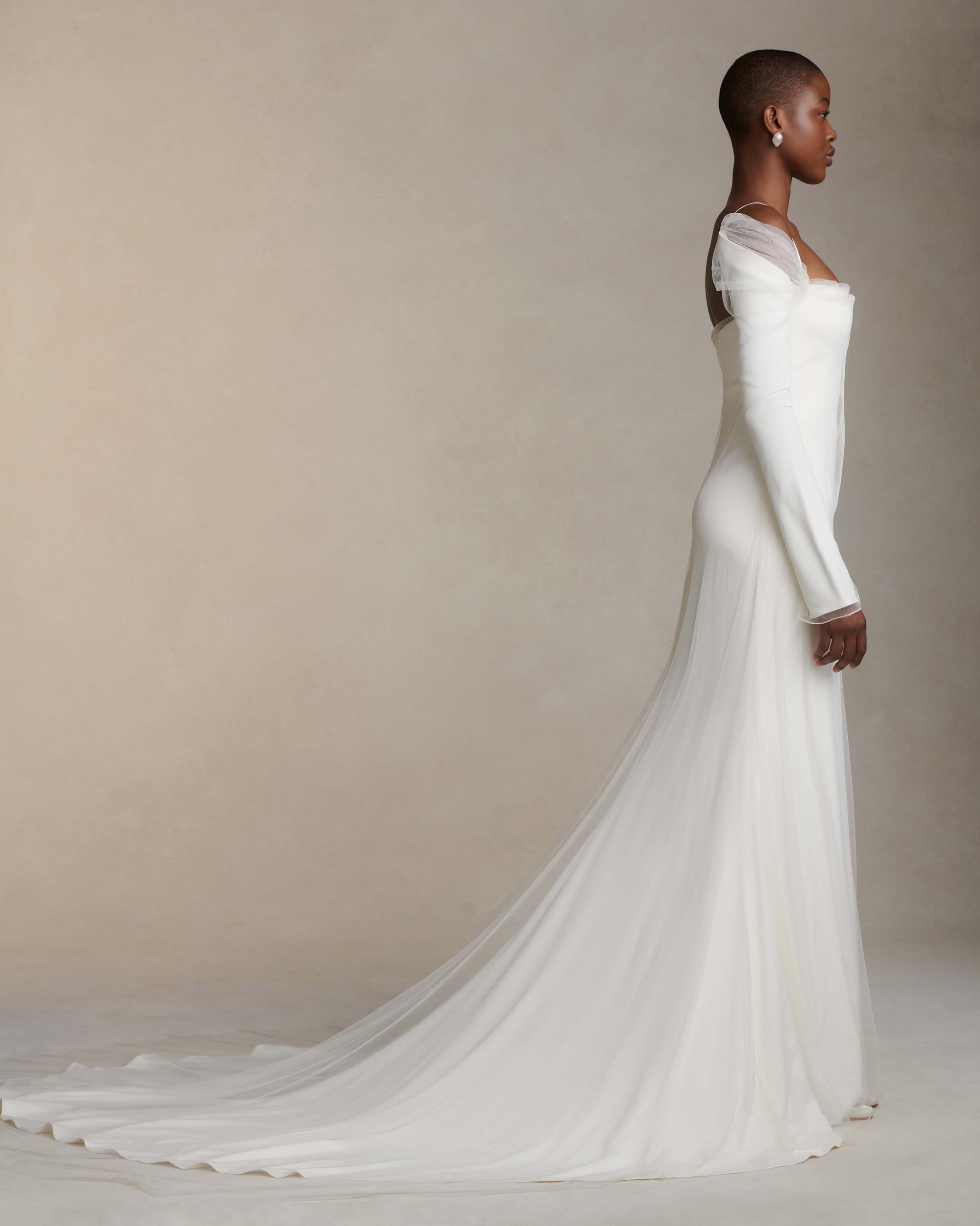 Stretch Cady off the shoulder gown with extended tulle overlay. Squared neckline accented by draped high tulle, long sleeves lightly bound to narrow satin ribbon straps. Center back external zipper and soft internal bustier with grosgrain waistband belt and three hook positions for support.