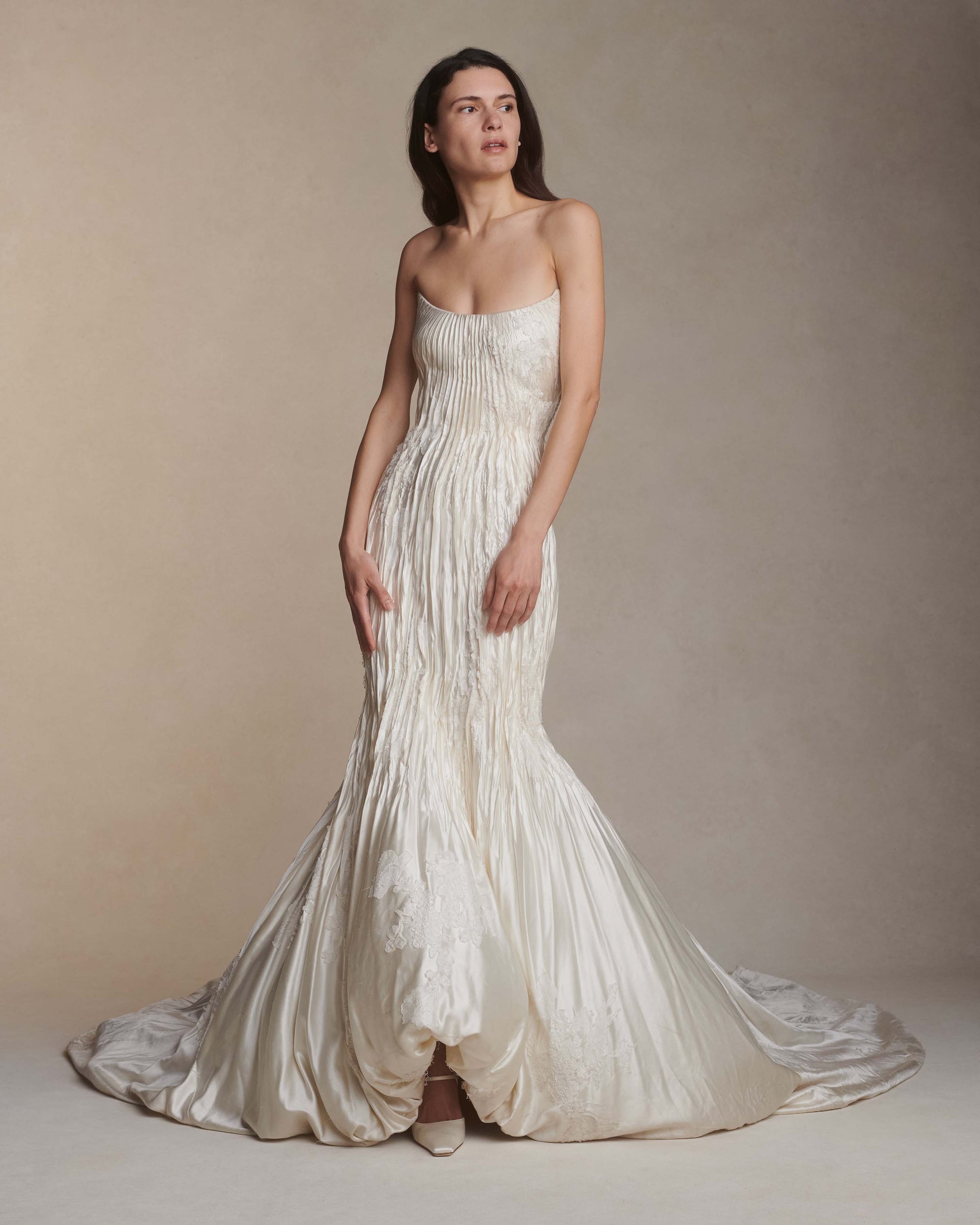 Silk wool hand crinkled gown fluted gown. Carved and sculpted strapless neckline. Hand appliquéd chantilly and corded lace motifs through body. Bubbled ankle grazing hem at front graduating into dramatic bubbled train. Soft internal corset with boning and center back zip closure.