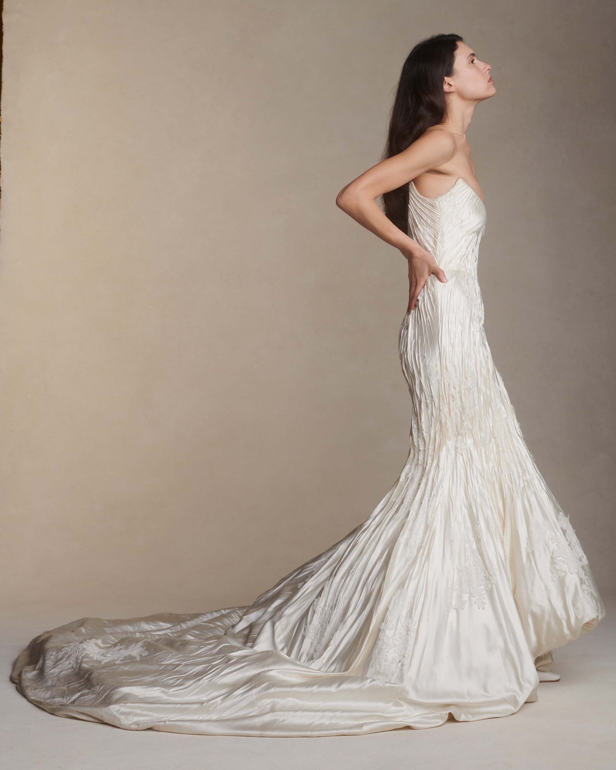 Silk wool hand crinkled gown fluted gown. Carved and sculpted strapless neckline. Hand appliquéd chantilly and corded lace motifs through body. Bubbled ankle grazing hem at front graduating into dramatic bubbled train. Soft internal corset with boning and center back zip closure.