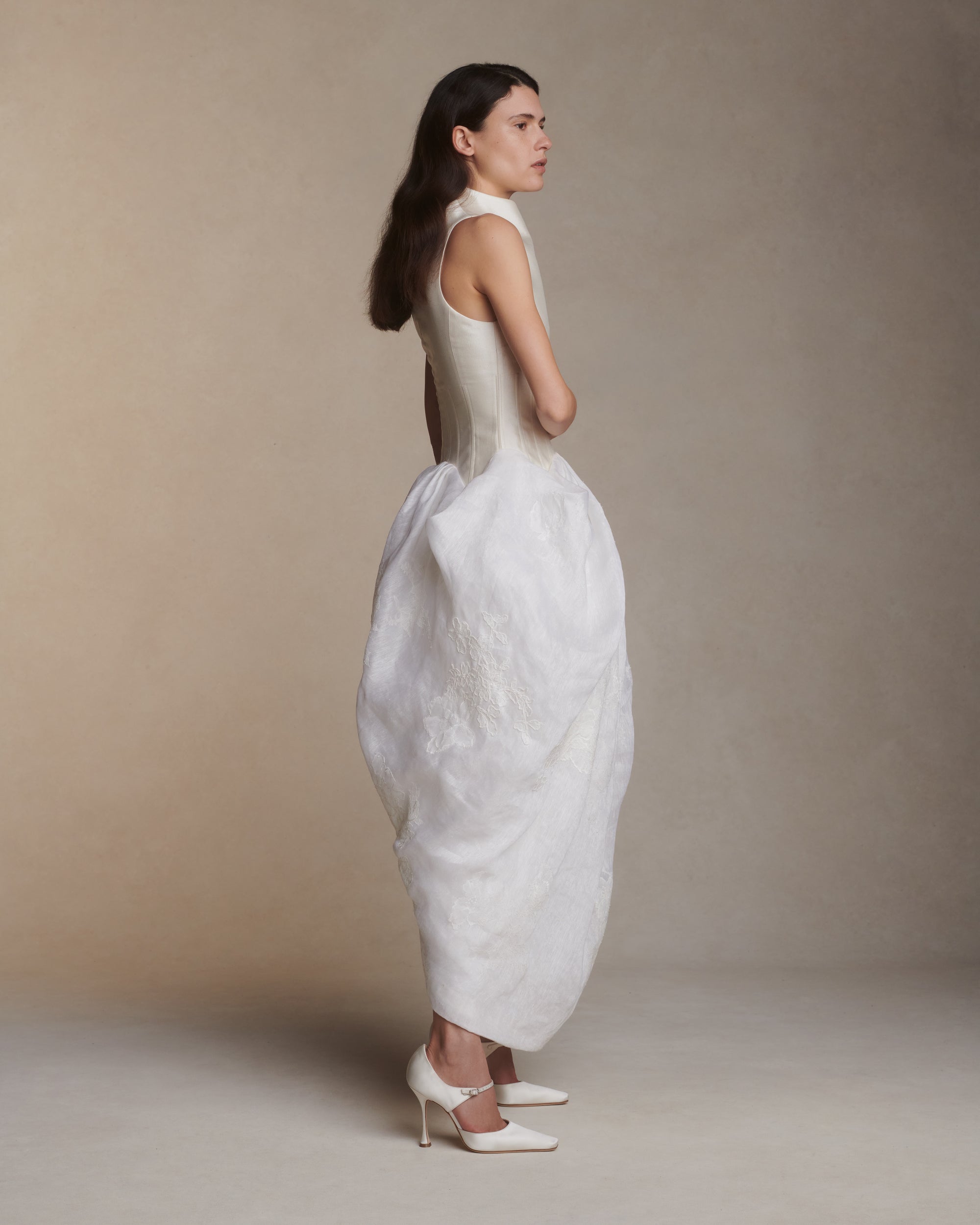 Silk wool twill and linen organza tea length gown. Structured boned bodice extends into dramatic sloped high neck that floats away from the body. Pleated and draped skirt is adorned with hand cut chantilly and corded lace appliques. Center back zip closure.