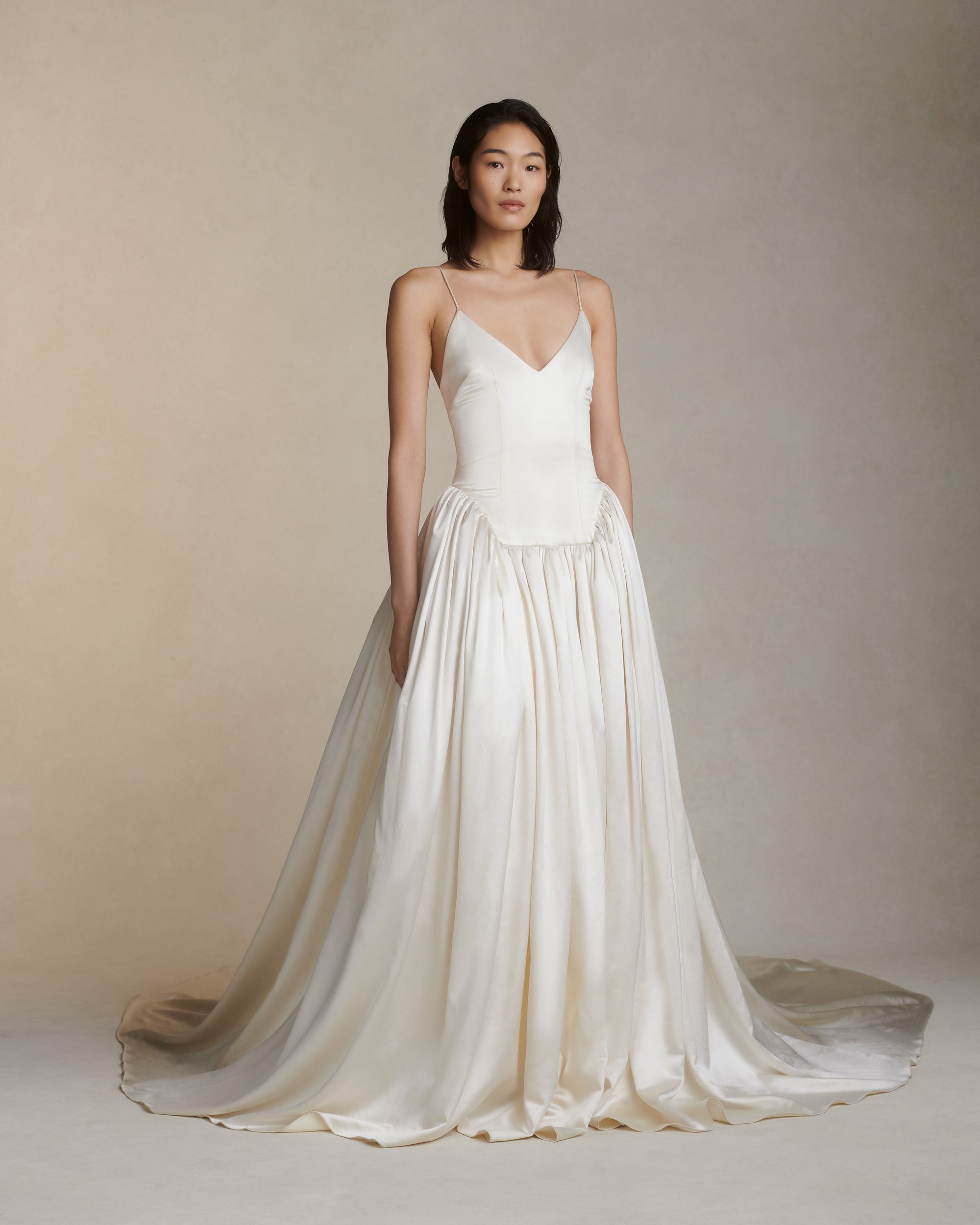 Deep V neck cathedral length gown with shirred, full skirt and elongated basque waist. Self lined, boned bodice for structure, and darts for shaping. Internal grosgrain waistband belt with three positions and center back zip. Fully lined soft skirt with exaggerated train, can can ruffle at hip for volume and structure.