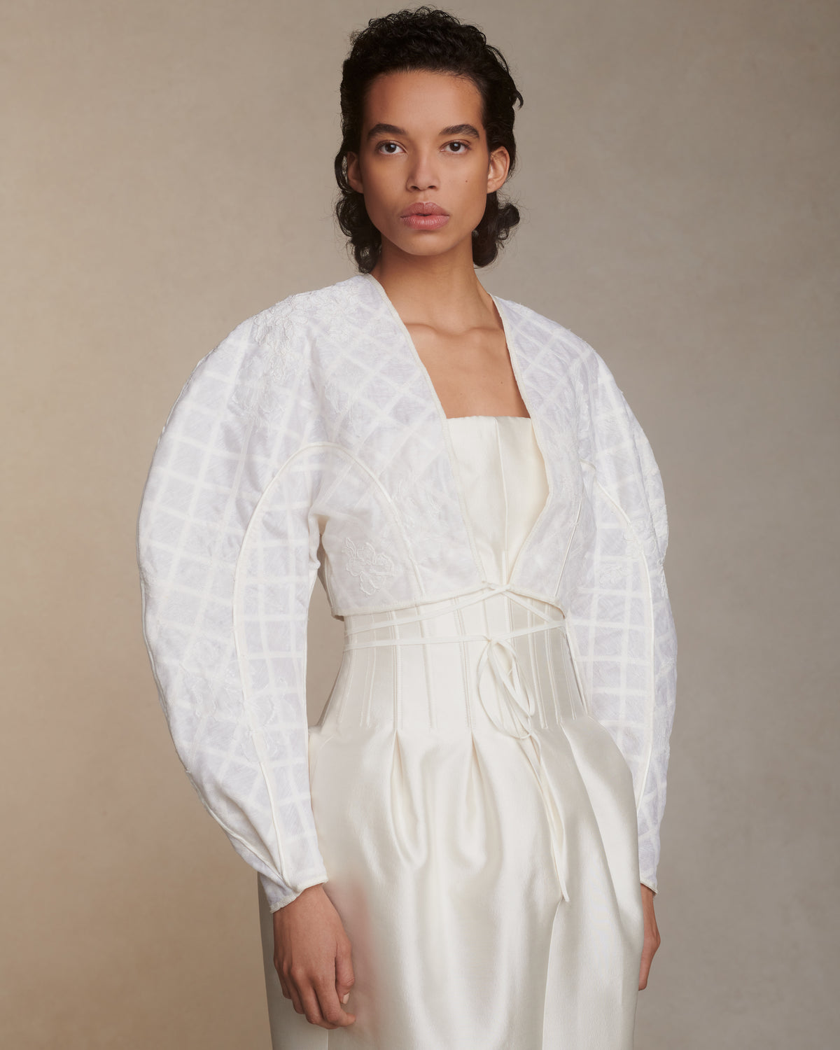 Sculptural linen organza cropped jacket framed with silk organza and grosgrain ribbon trellis detail. Hand cut and chantilly lace appliqued throughout. Satin piping detail and long grosgrain ribbon tie detail at center front. Covered button and loop closure at sleeve opening.Styled with Audra gown