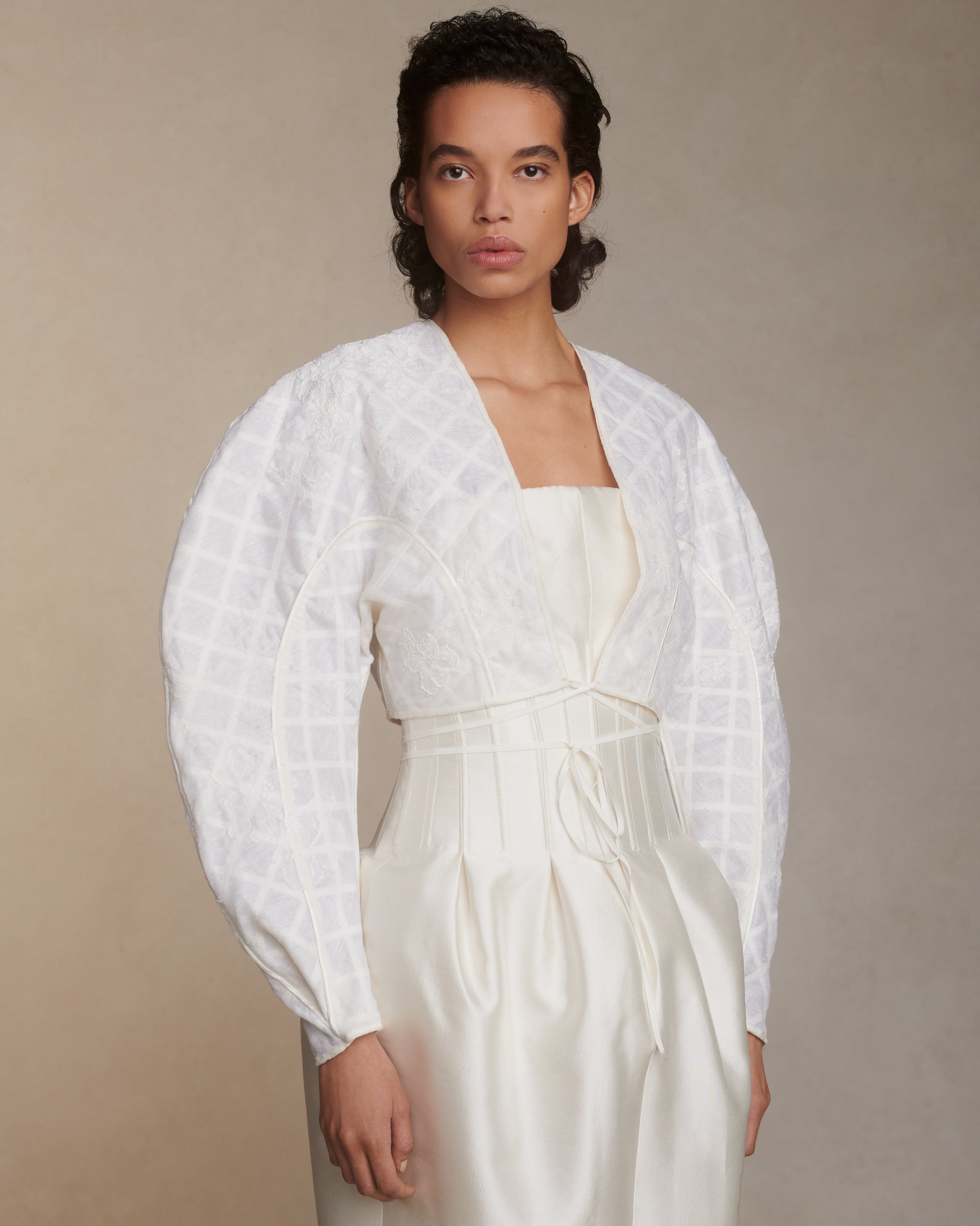 Sculptural linen organza cropped jacket framed with silk organza and grosgrain ribbon trellis detail. Hand cut and chantilly lace appliqued throughout. Satin piping detail and long grosgrain ribbon tie detail at center front. Covered button and loop closure at sleeve opening.Styled with Audra gown