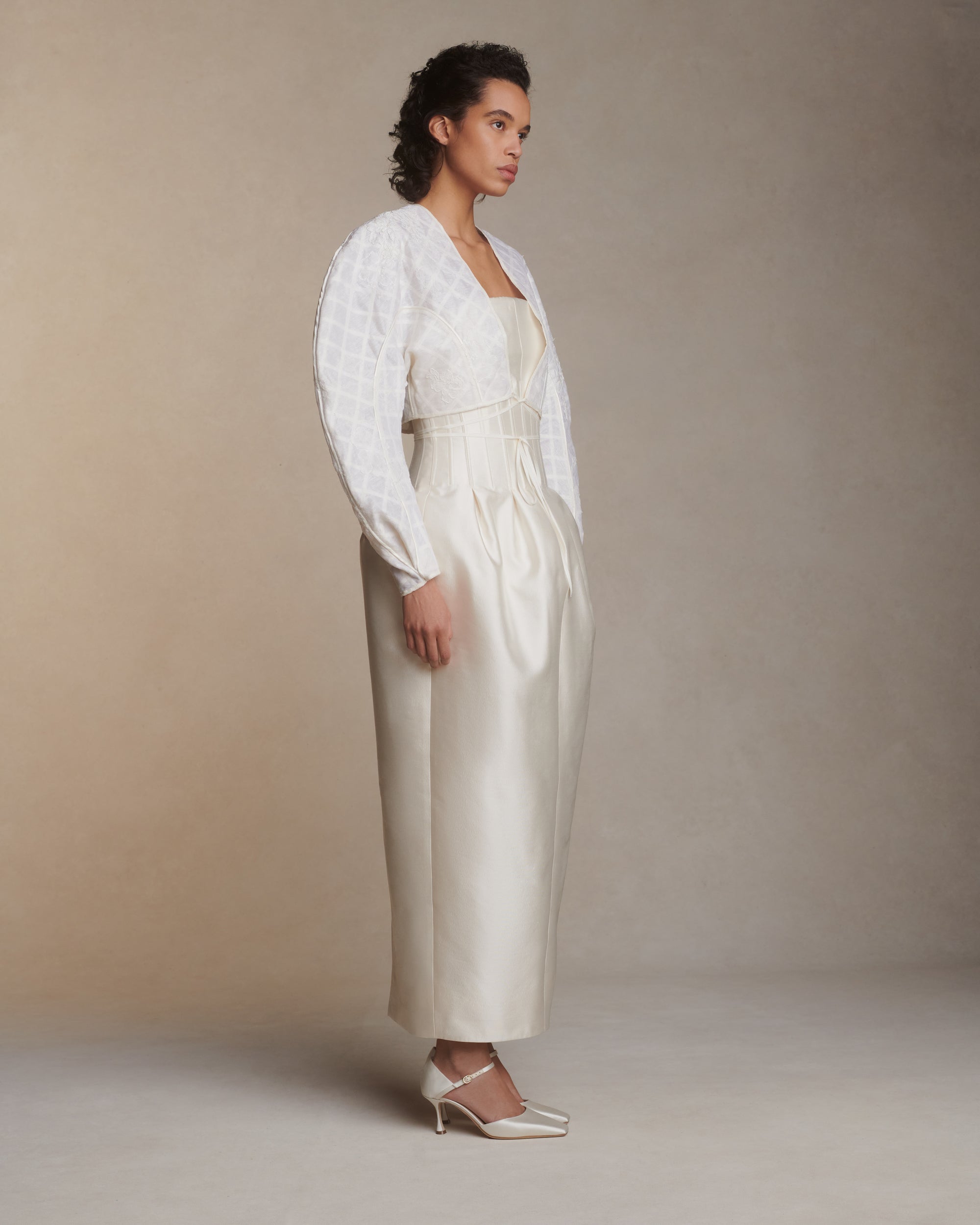 Sculptural linen organza cropped jacket framed with silk organza and grosgrain ribbon trellis detail. Hand cut and chantilly lace appliqued throughout. Satin piping detail and long grosgrain ribbon tie detail at center front. Covered button and loop closure at sleeve opening.Styled with Audra gown