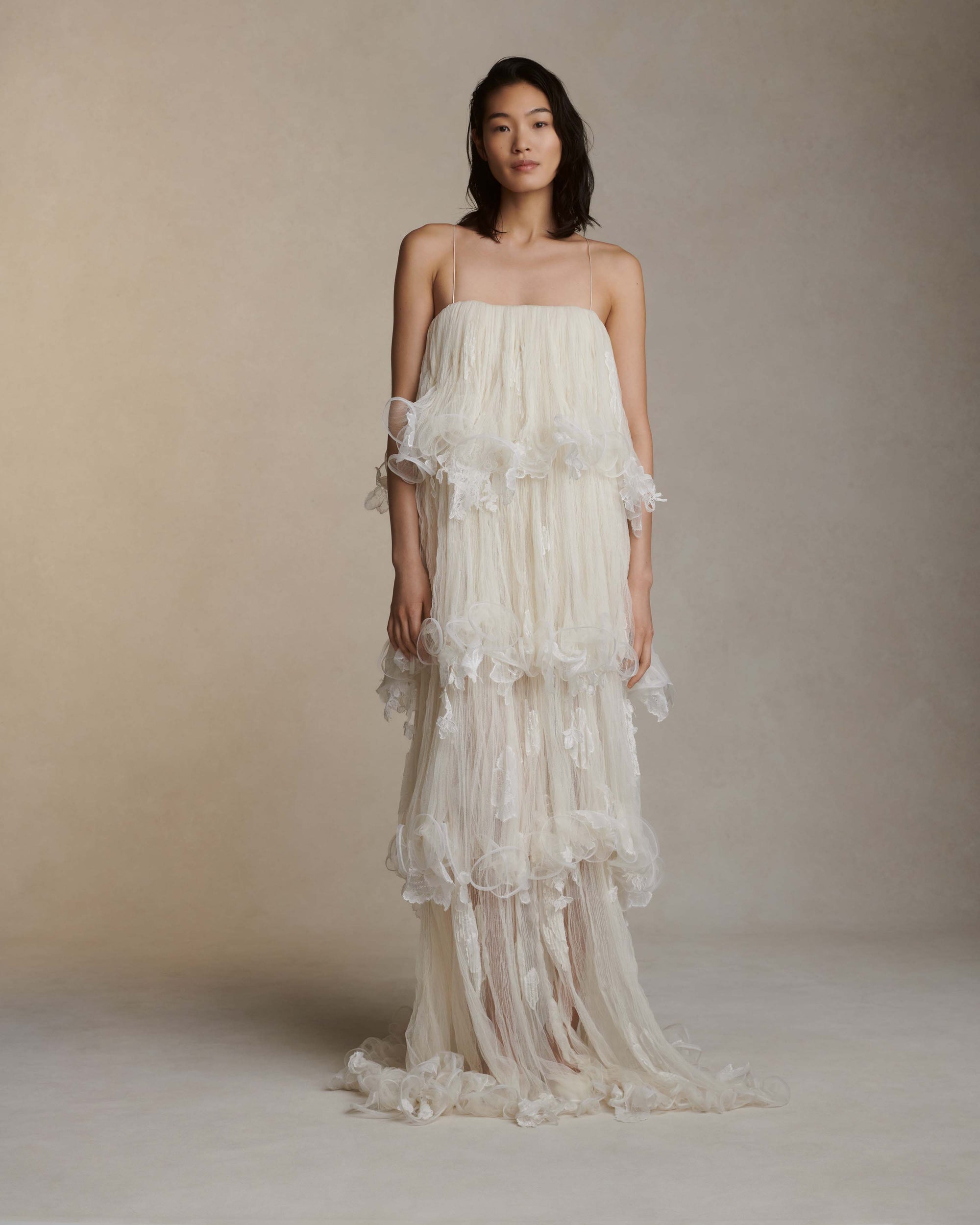 Tiered tulle gown with hand appliquéd chantilly and corded lace. Four layered tulle body pronounced by delicate horsehair edge. Below knee lining for slight transparency at calf. Soft square neckline with spaghetti strap. Free of cups and boning and finished with center back zip close.