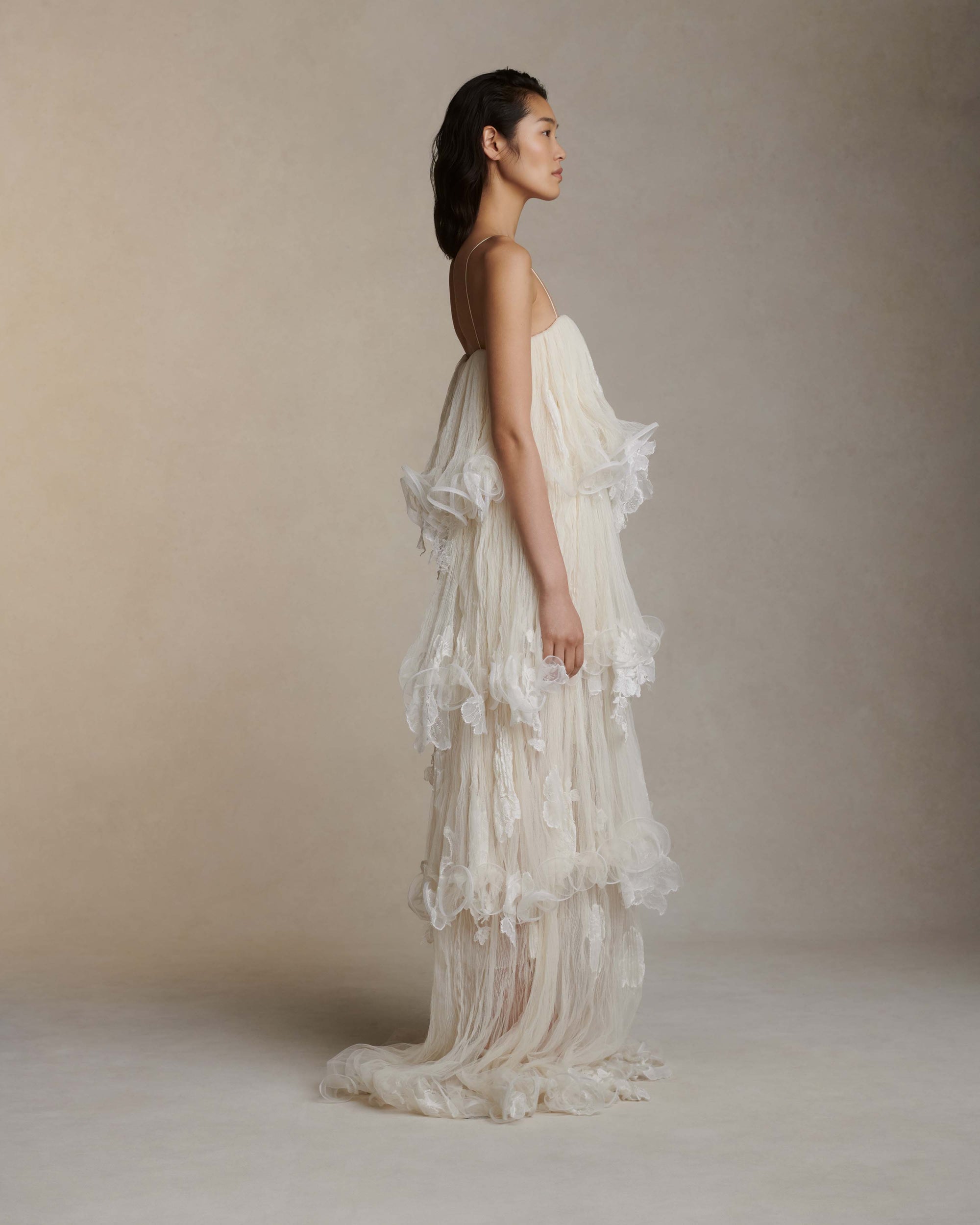 Tiered tulle gown with hand appliquéd chantilly and corded lace. Four layered tulle body pronounced by delicate horsehair edge. Below knee lining for slight transparency at calf. Soft square neckline with spaghetti strap. Free of cups and boning and finished with center back zip close.