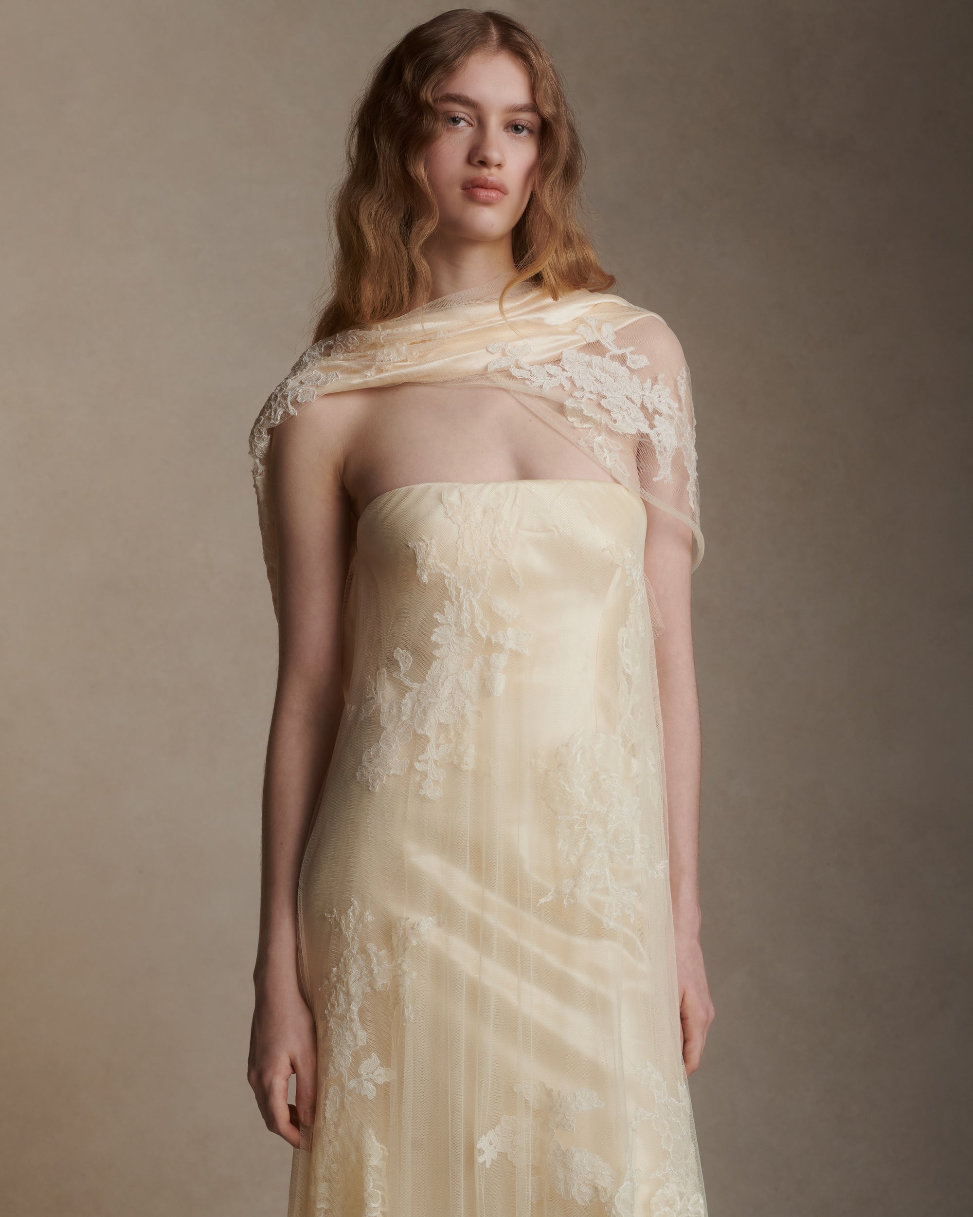 Strapless satin gown with soft tulle overlay decorated in hand cut corded and chantilly lace appliques. Asymmetric drape wraps gracefully across neck and shoulders falling into a scarf drape detail at low back. Bubble hem releases into train at back. Boned internal bustier with soft cups and center back zip closure.