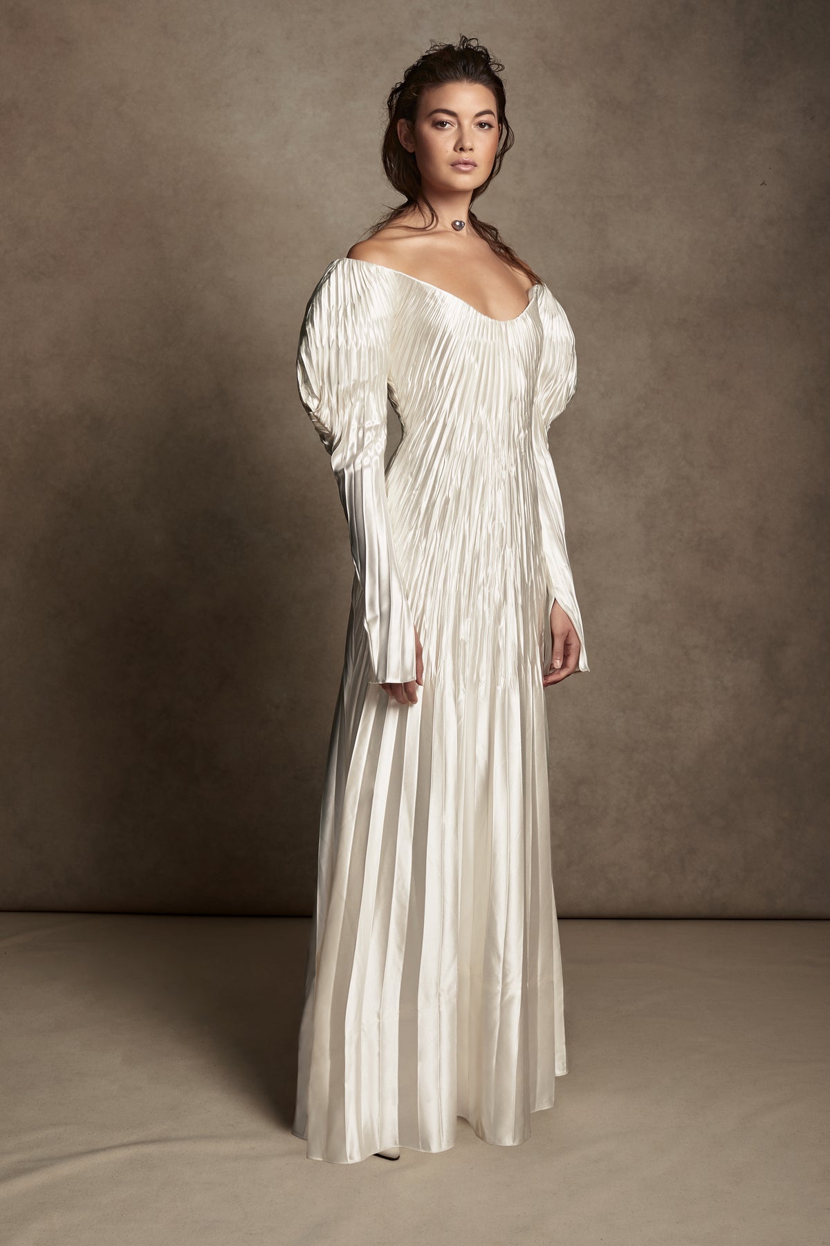 Pleated silk wool, exaggerated off the shoulder, floor length gown. Stiff neckline facing stay for structure and positioning of bodice framed by full length sleeves and 5 inch slit at inner wrist. Full internal power mesh corset with boning and internal and external center back zippers for security.Styled with Flaired Baroque Pearl Choker