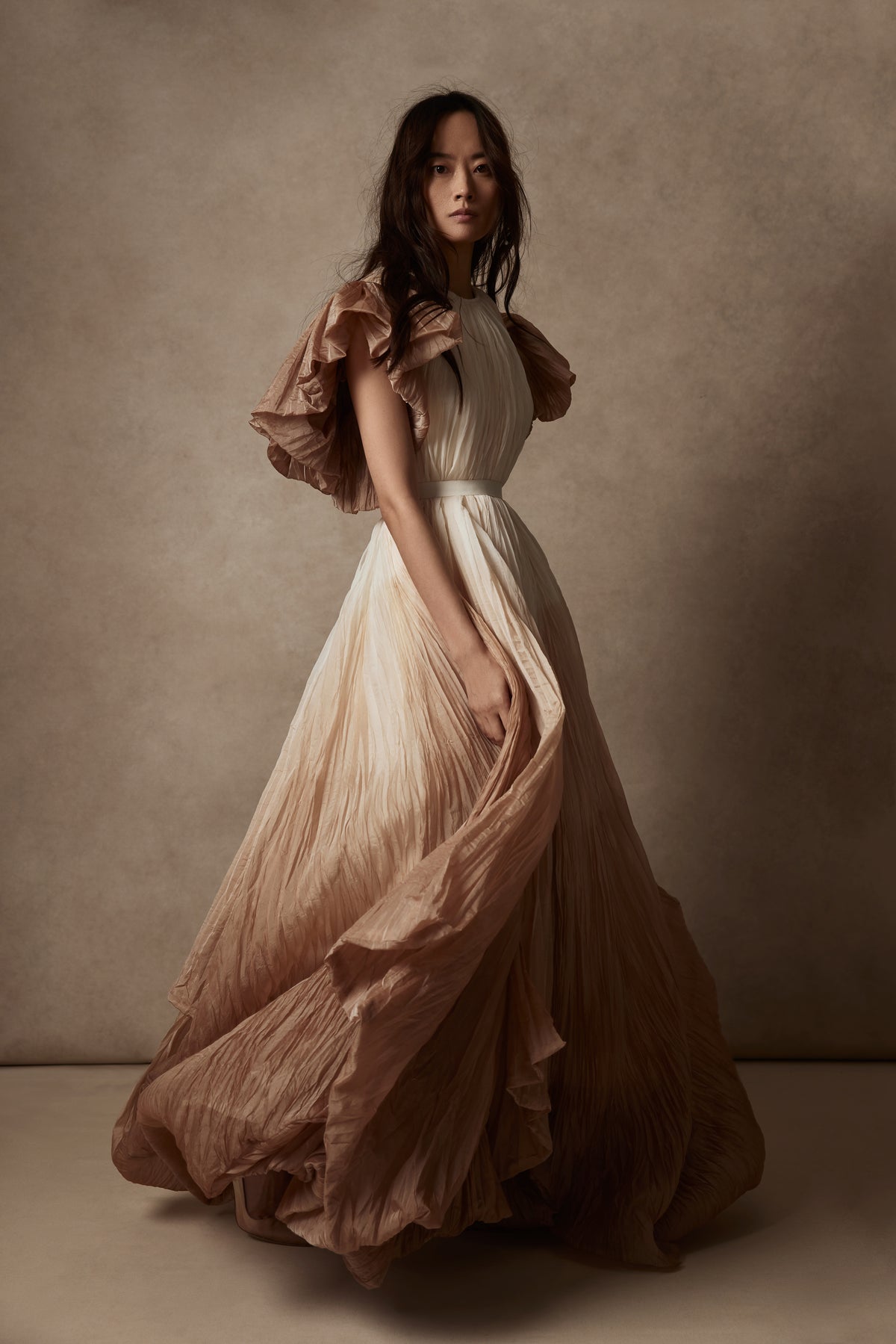 Dip dyed pleated paper organza gown with bubble hem and voluminous short ruffled sleeve. Center back zipper and external grosgrain belt atop soft tulle lined internal bustier. Crew neckline with button close at back of neck creating triangular shaped open back. Available in ivory upon request.