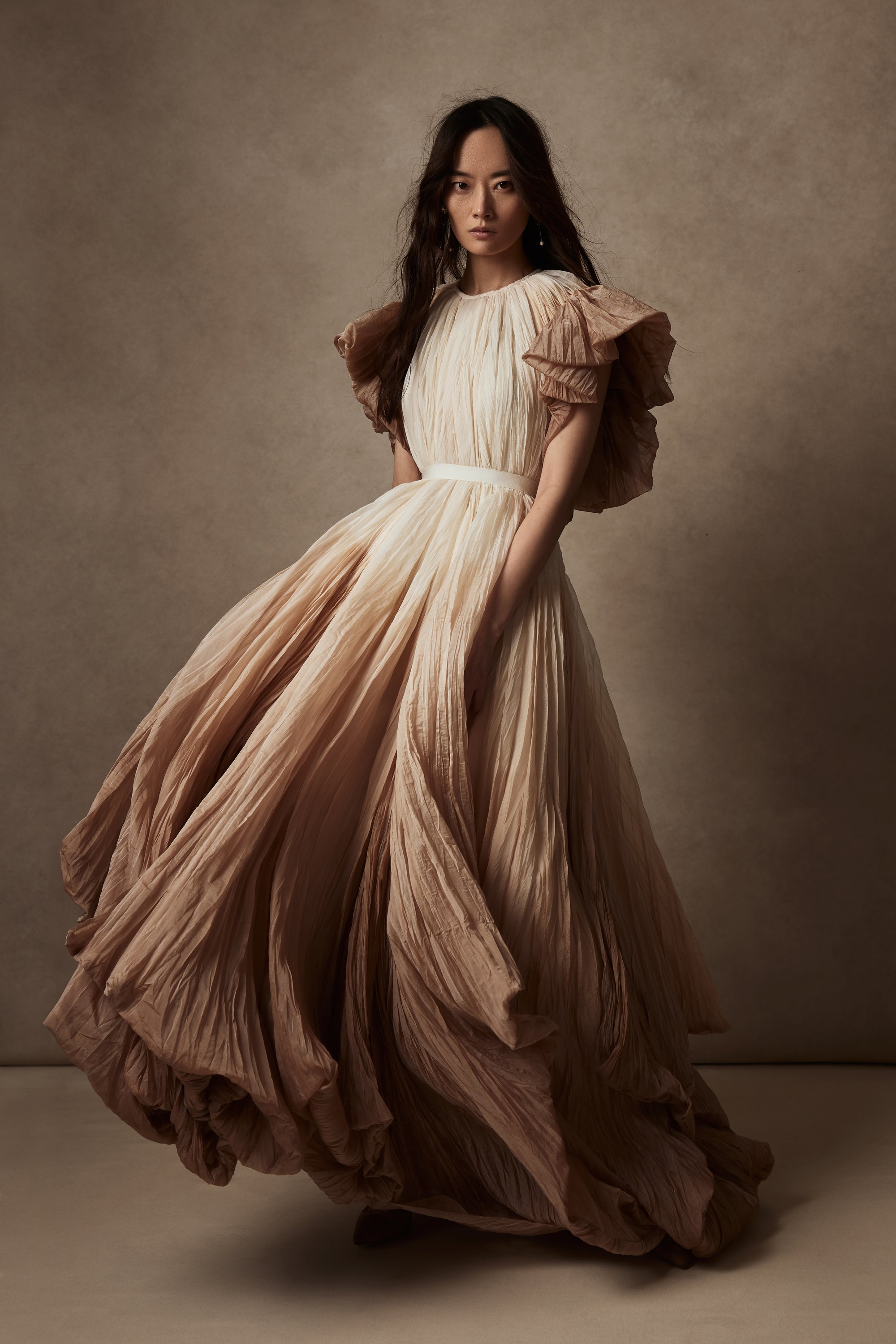 Dip dyed pleated paper organza gown with bubble hem and voluminous short ruffled sleeve. Center back zipper and external grosgrain belt atop soft tulle lined internal bustier. Crew neckline with button close at back of neck creating triangular shaped open back. Available in ivory upon request.