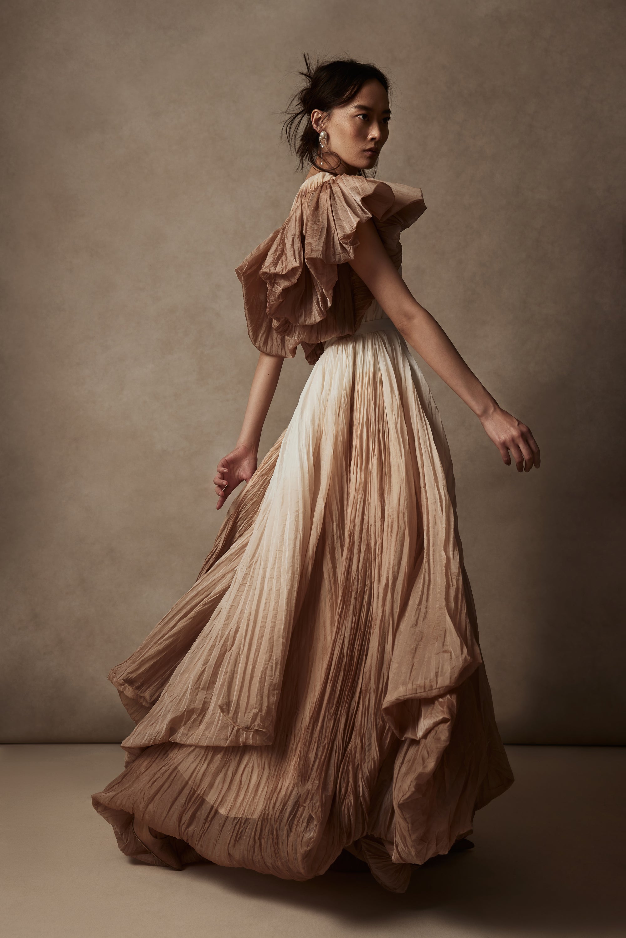 Dip dyed pleated paper organza gown with bubble hem and voluminous short ruffled sleeve. Center back zipper and external grosgrain belt atop soft tulle lined internal bustier. Crew neckline with button close at back of neck creating triangular shaped open back. Available in ivory upon request.